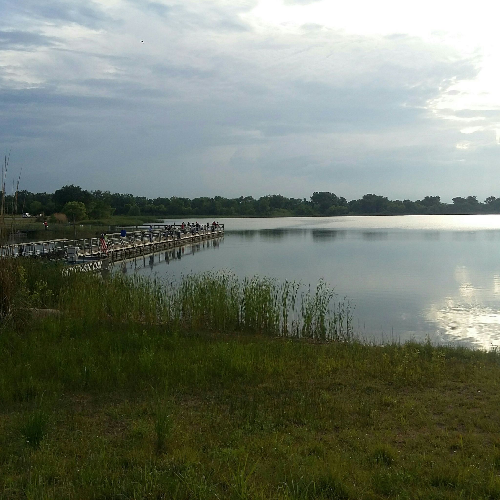 Long pier with people extending into a calm lake under cloudy sky. - West River Trail