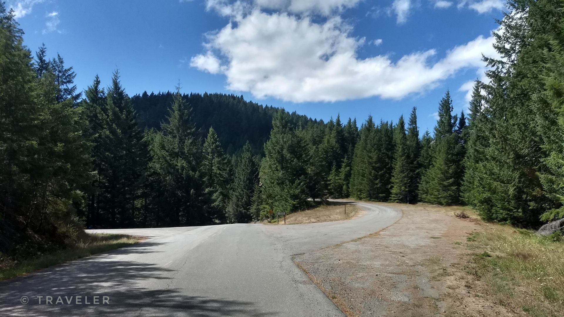 Paved road winding through a dense evergreen forest under a partly cloudy sky. - Powers to Agness Saddle