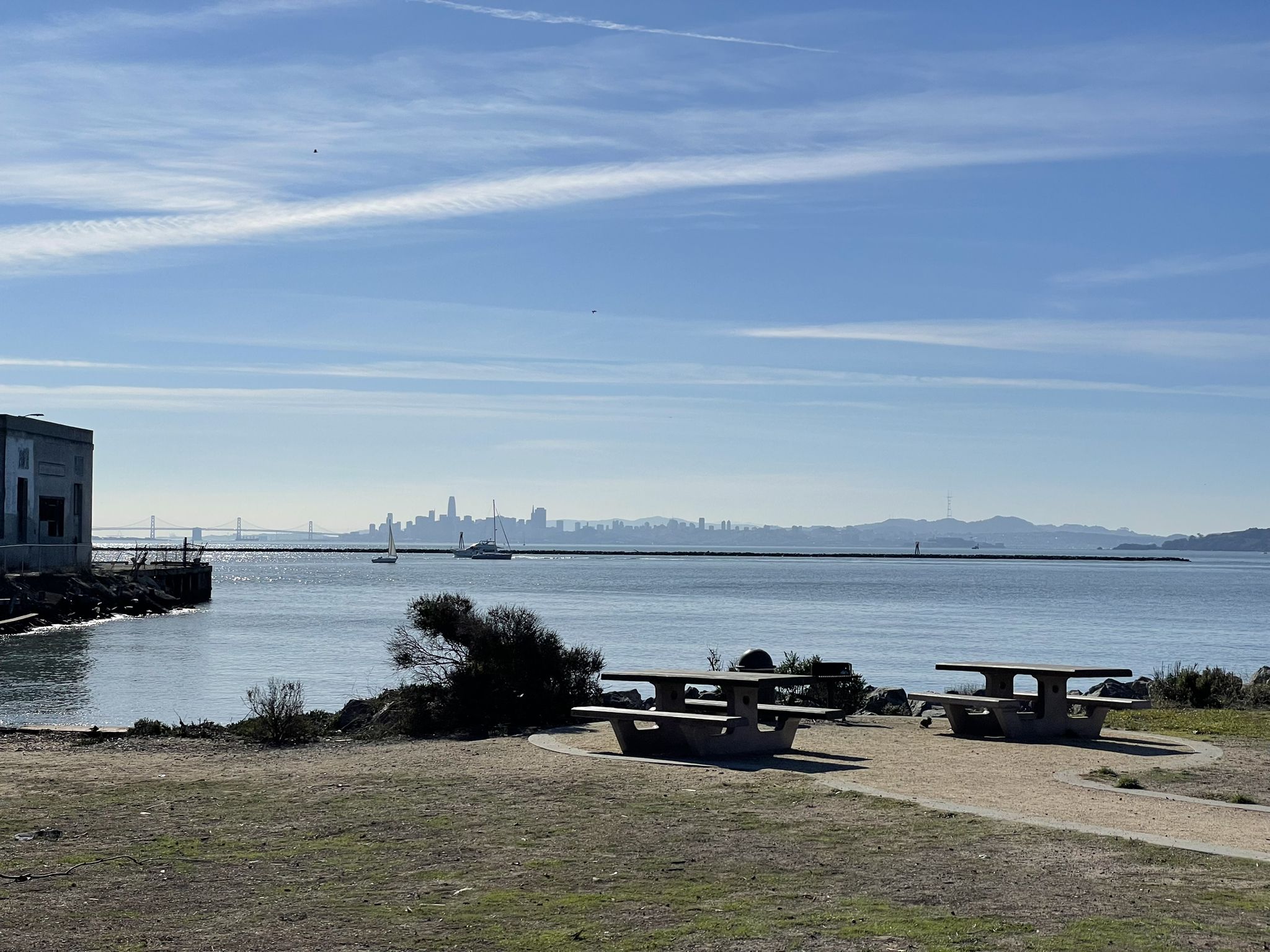 Picnic tables by calm water with a distant city skyline under blue sky. - Ferry Point Loop and Shipyard 3 Trail