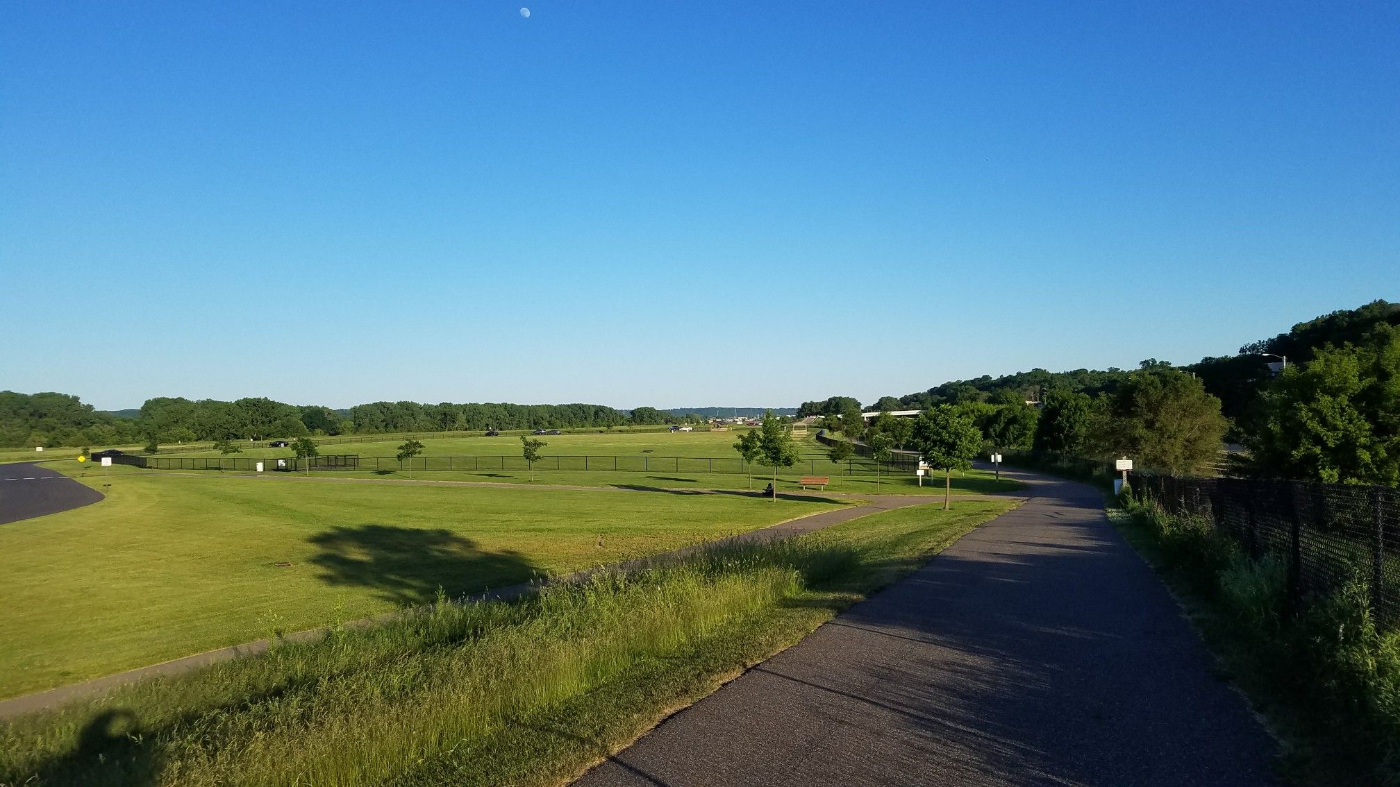Paved path winding through vast green fields under a clear blue sky. - Mississippi River Greenway