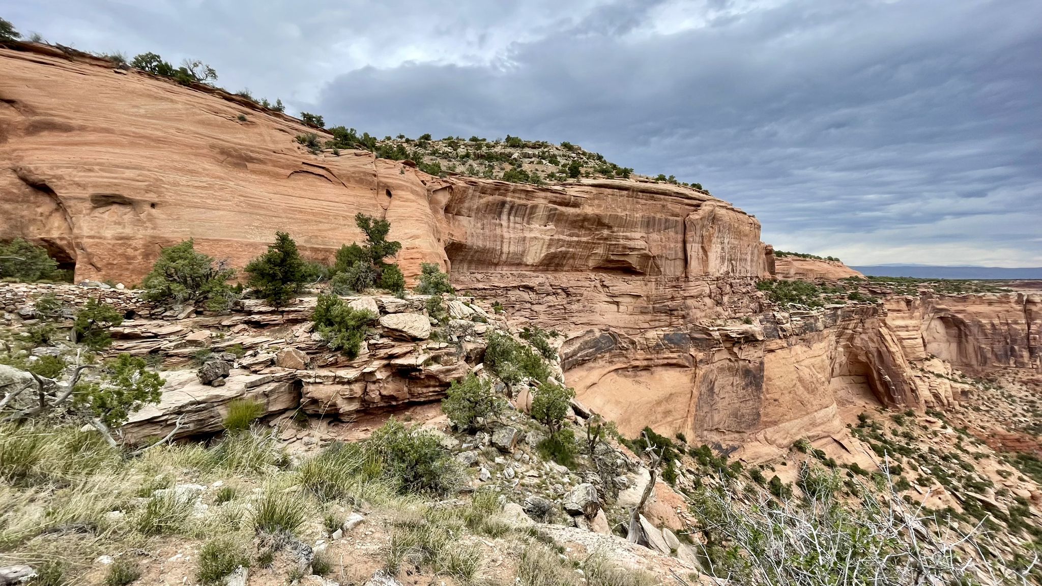 Red rock cliffs and sparse green vegetation under a cloudy sky. - Coke Ovens Trail