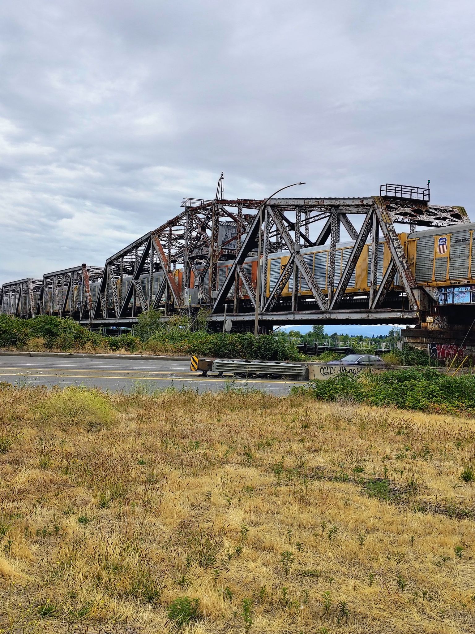 A long freight train crosses a rusty metal bridge under a cloudy sky. - Roadbike loop from North Lombard Transit Center