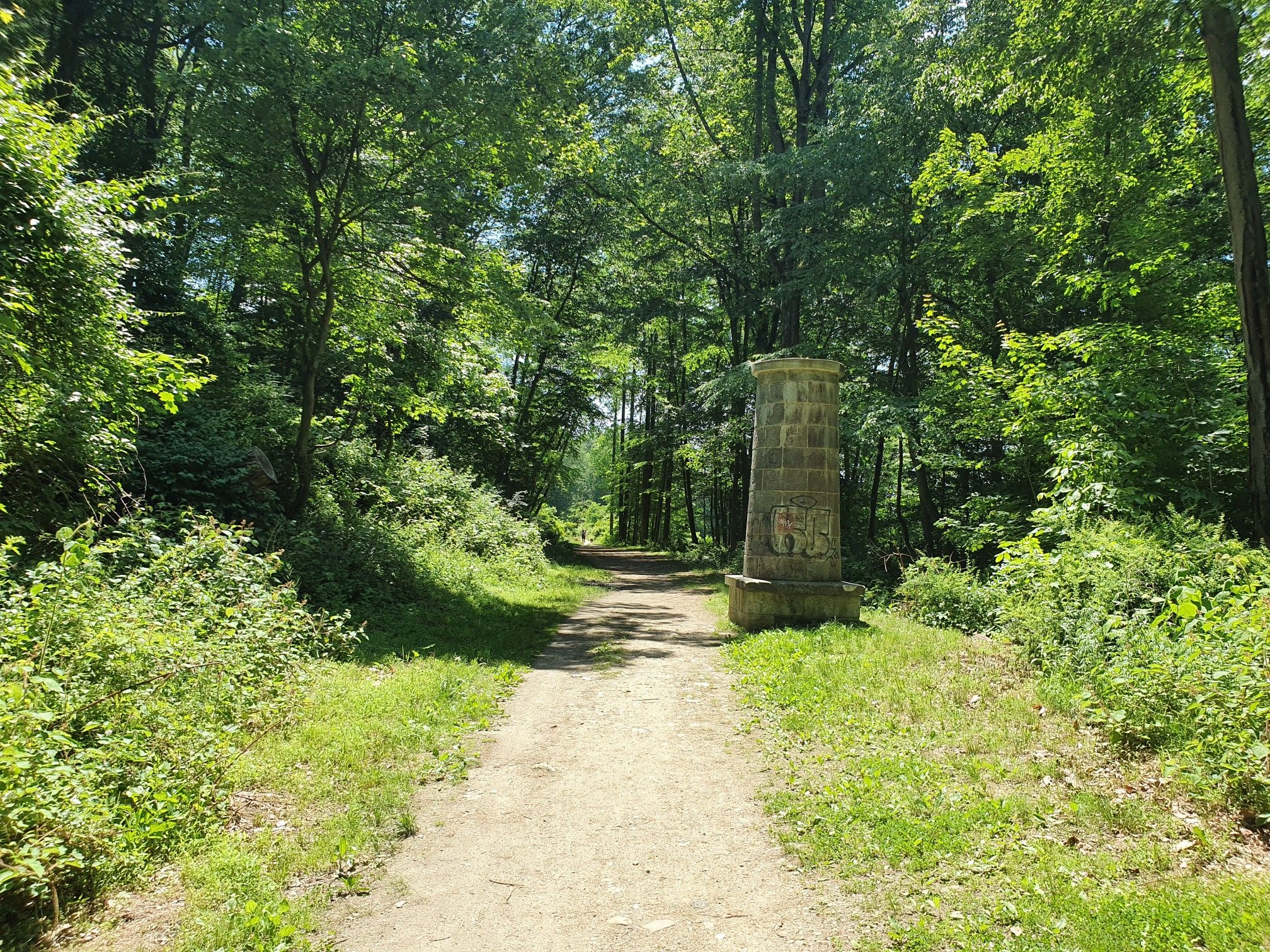 Sunny dirt path through a green forest, past an old stone pillar. - Old Croton Aqueduct Loop