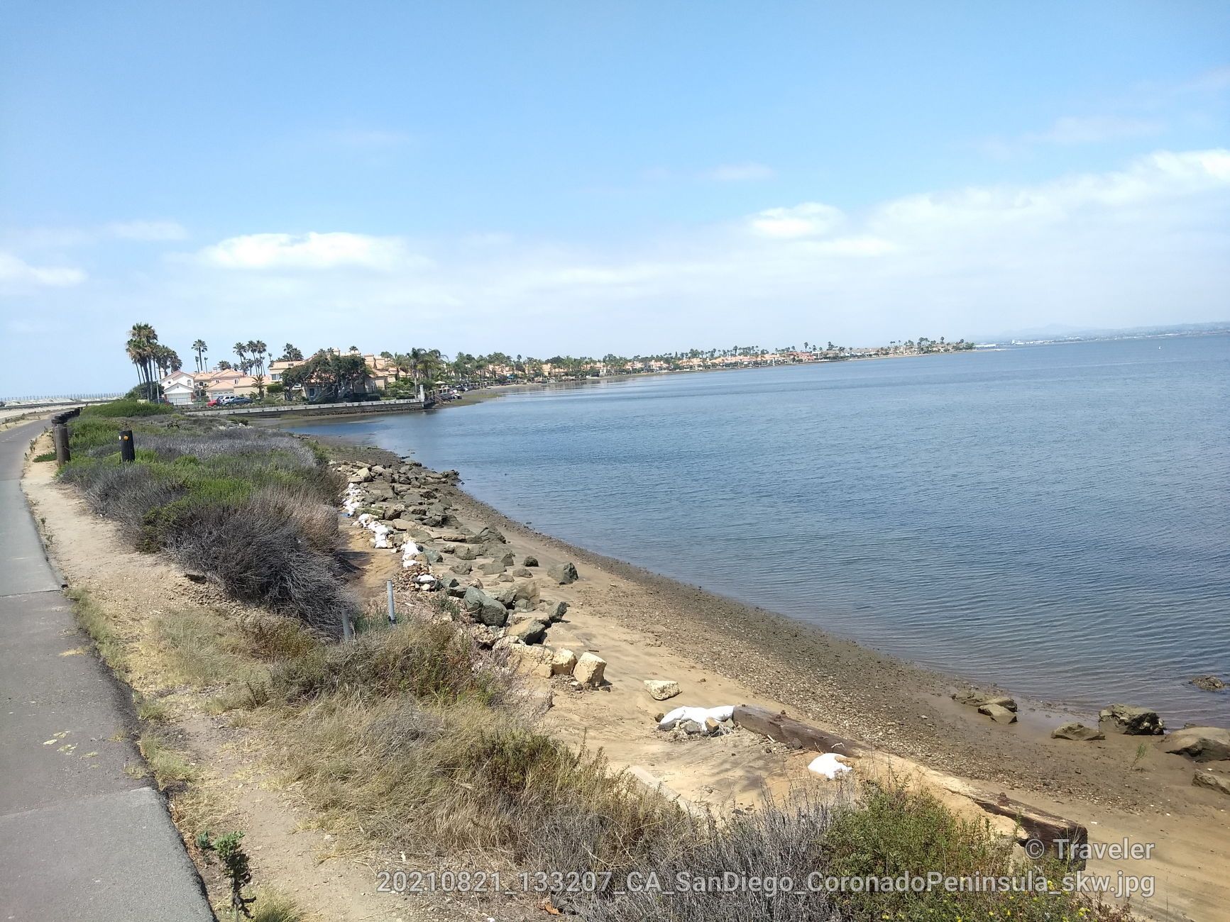 Paved path along a rocky shore, calm blue water, and distant buildings. - Coronado Bayshore Bikeway – View of San Diego Bay loop from San Diego