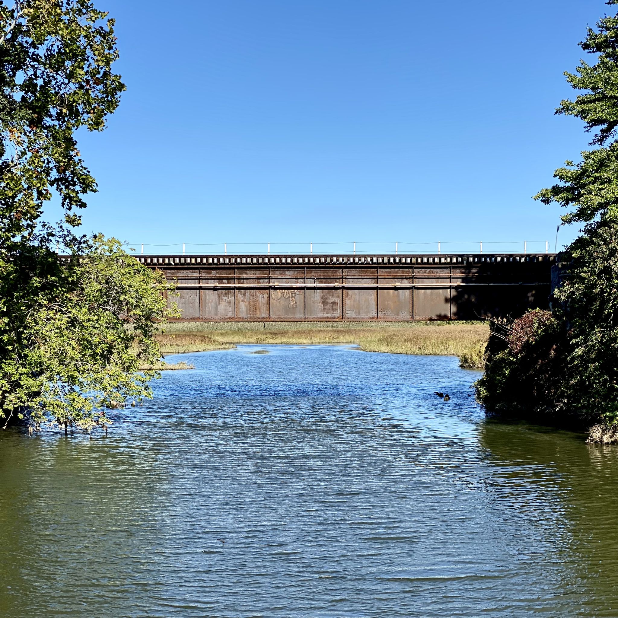 A large metal bridge spans a narrow waterway under a clear blue sky. - East Coast Greenway and 9/11 Trail Loop