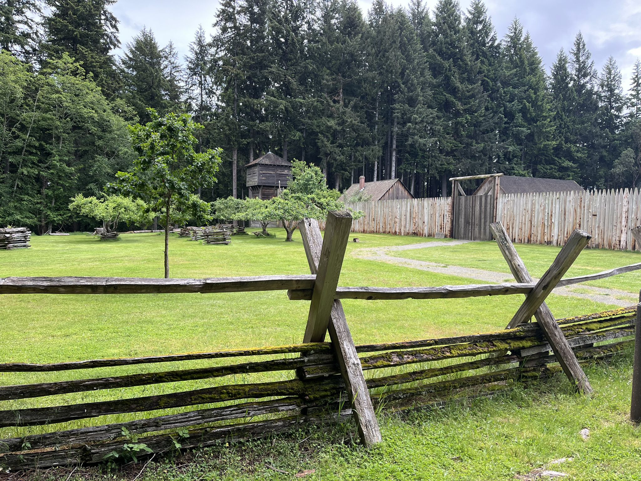 Wooden split-rail fence in front of a grassy field and fort structures. - Fort Nisqually Loop
