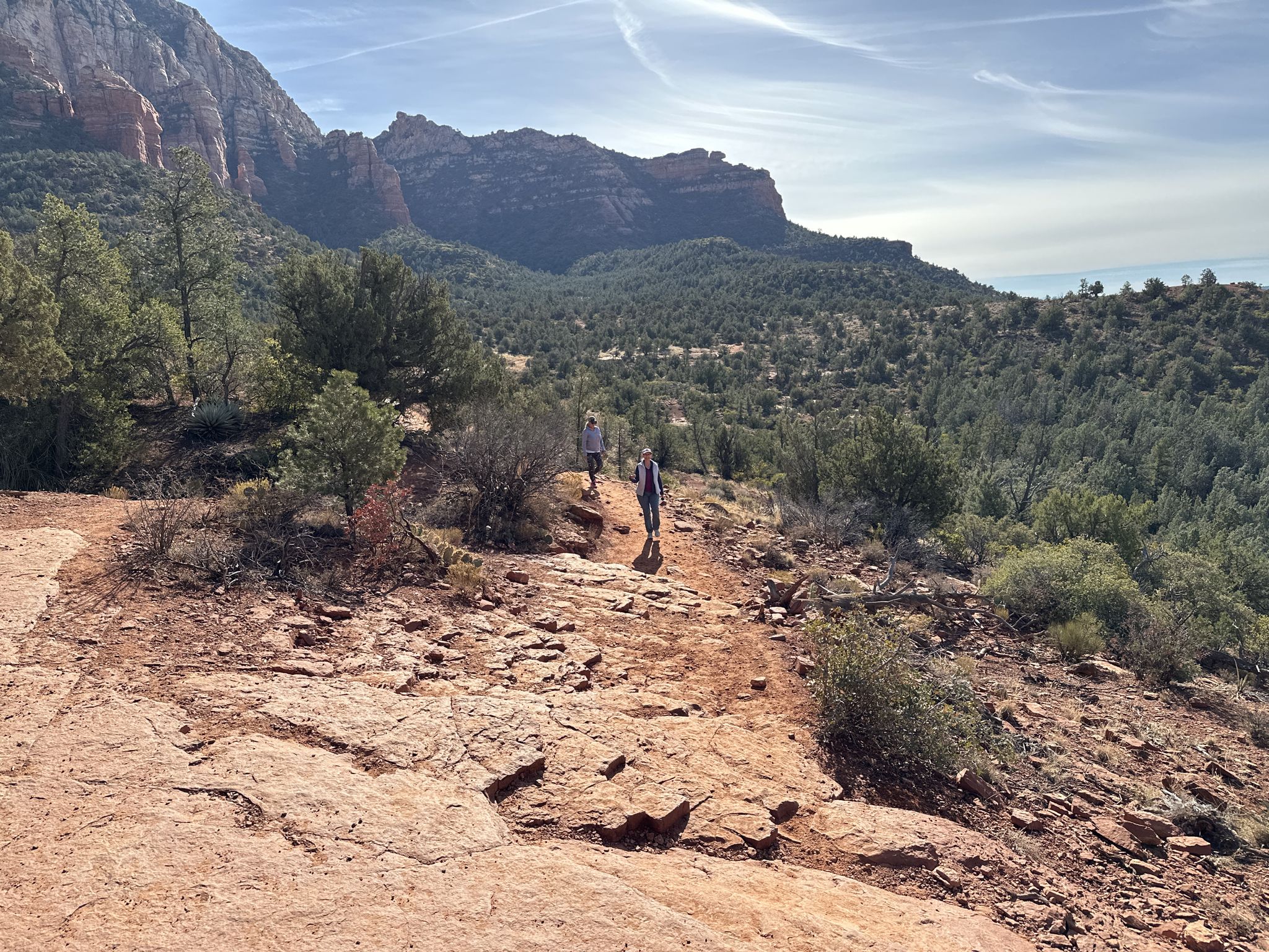 Two hikers on a rocky red trail with distant mountains and green trees. - Mescal Mountain Loop