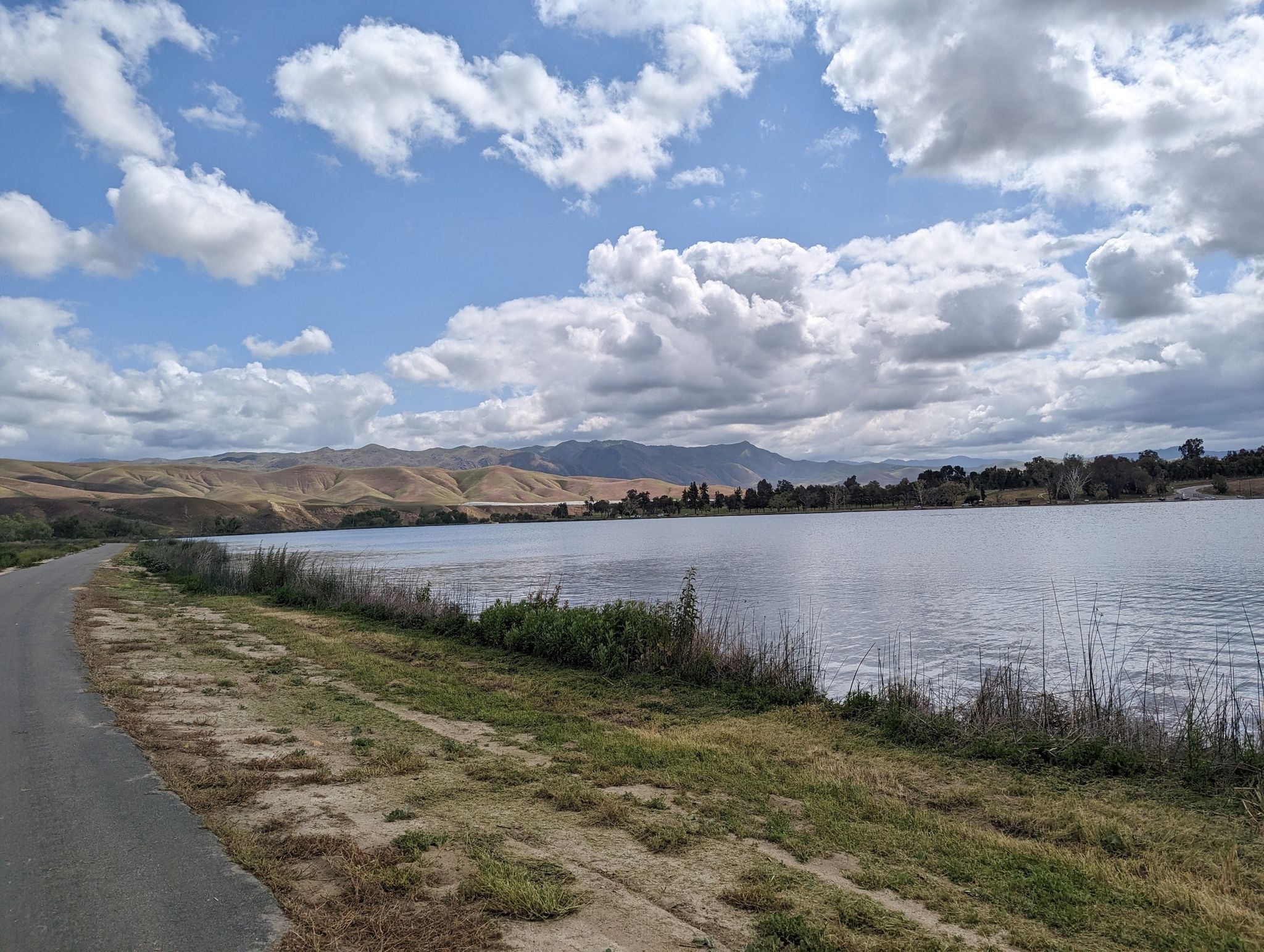 A paved road next to a lake with distant hills under a cloudy sky. - Ming Lake Loop