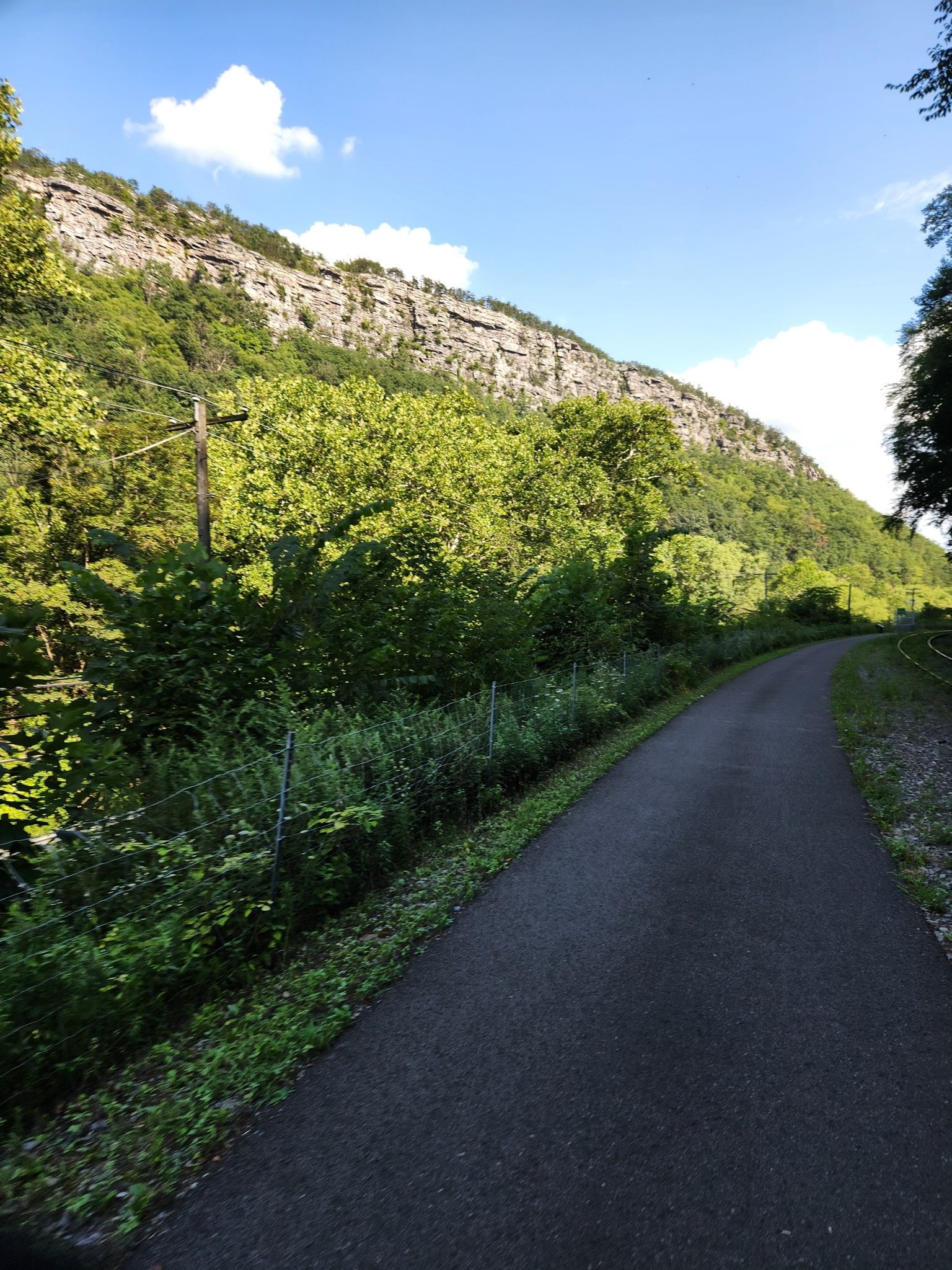 Paved path winds beside a steep, rocky, tree-covered mountain on a sunny day. - Roadbike loop from Cumberland