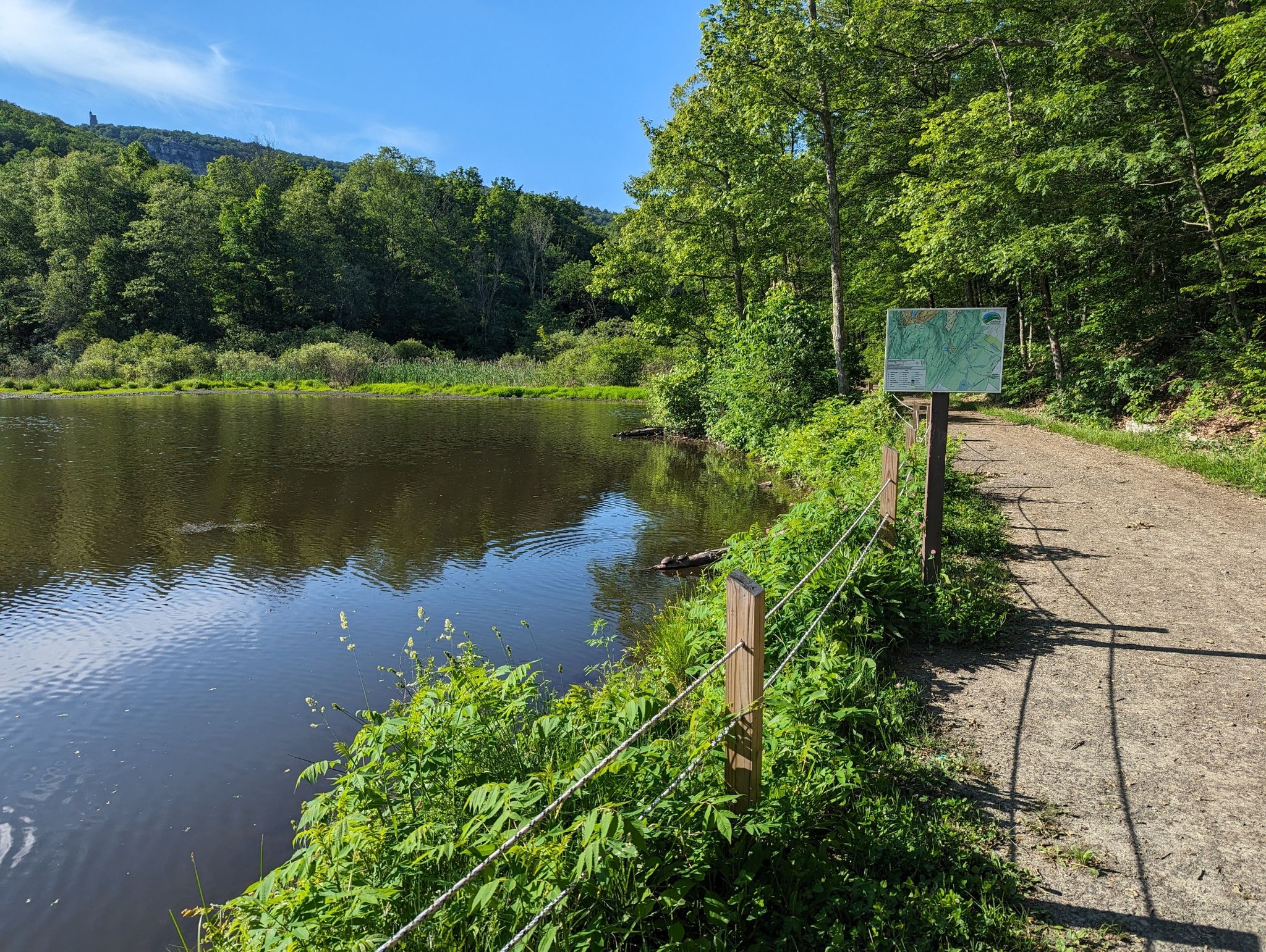 Sunny dirt path beside a calm lake with lush green trees and a sign. - Lower Duck Pond Road