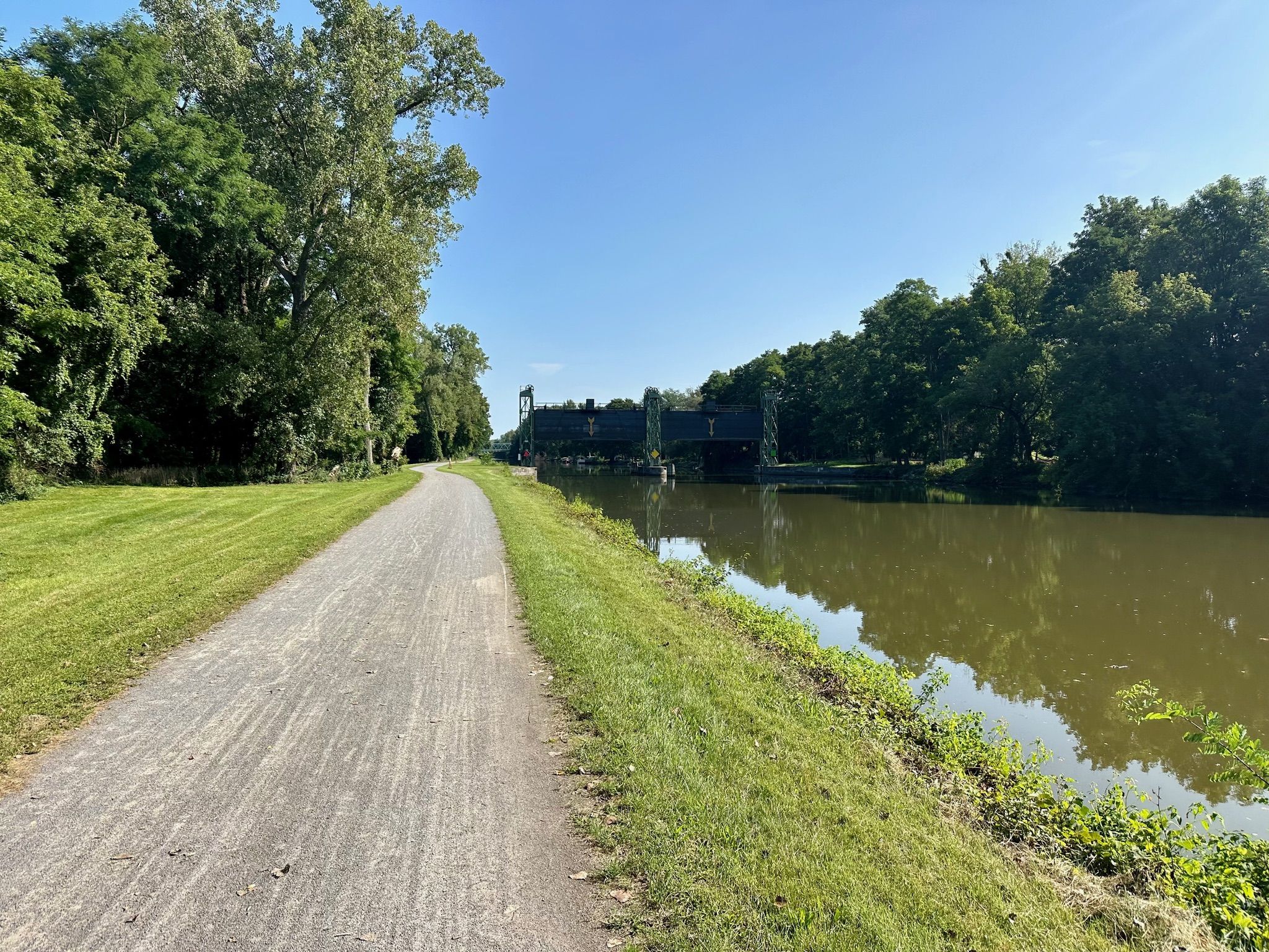 A dirt path next to a river with a large bridge in the distance, sunny. - McCoord Woods loop from Perinton
