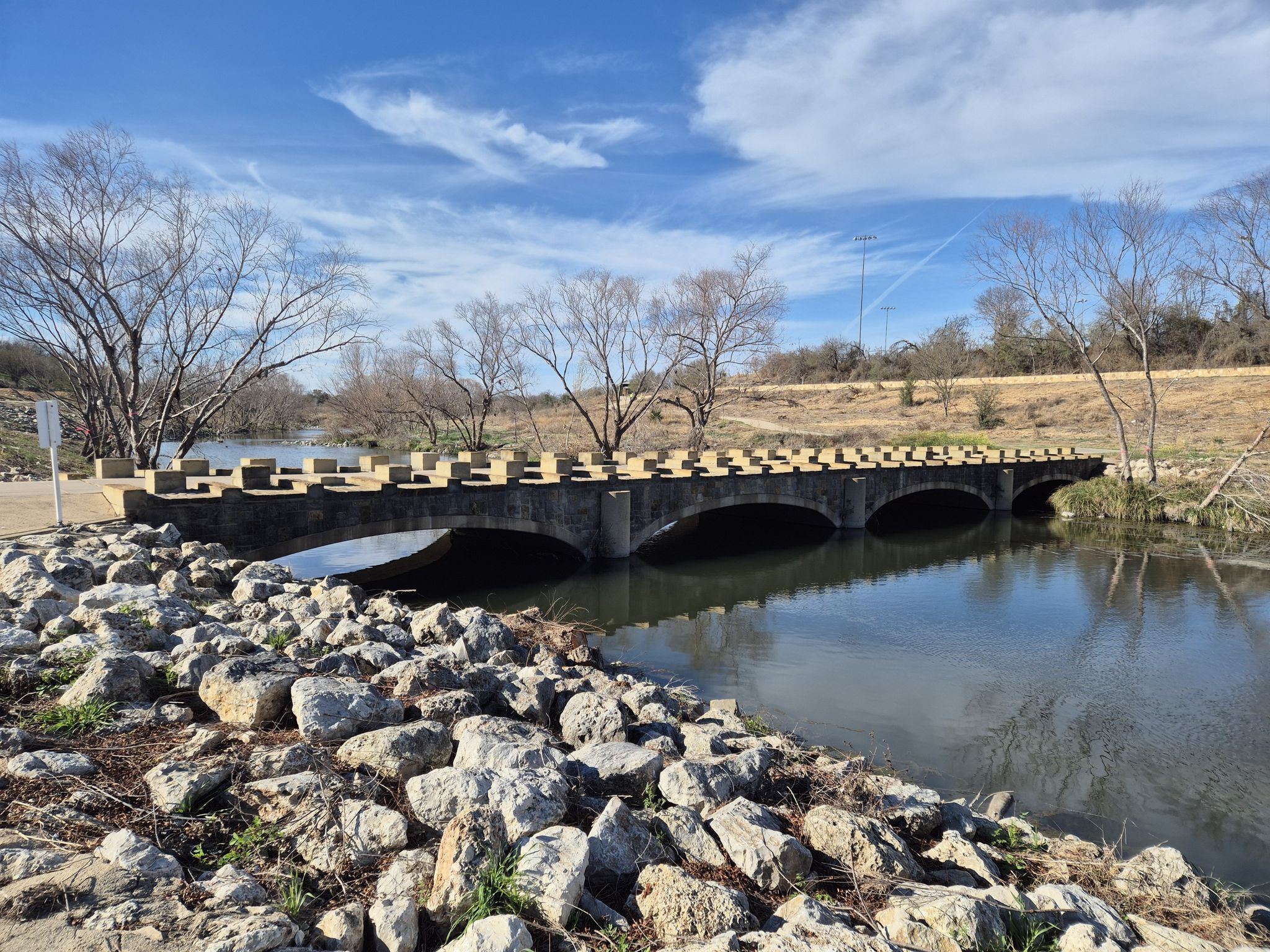 Stone bridge over a calm river, surrounded by bare trees, bright sky. - River Walk Mission Reach