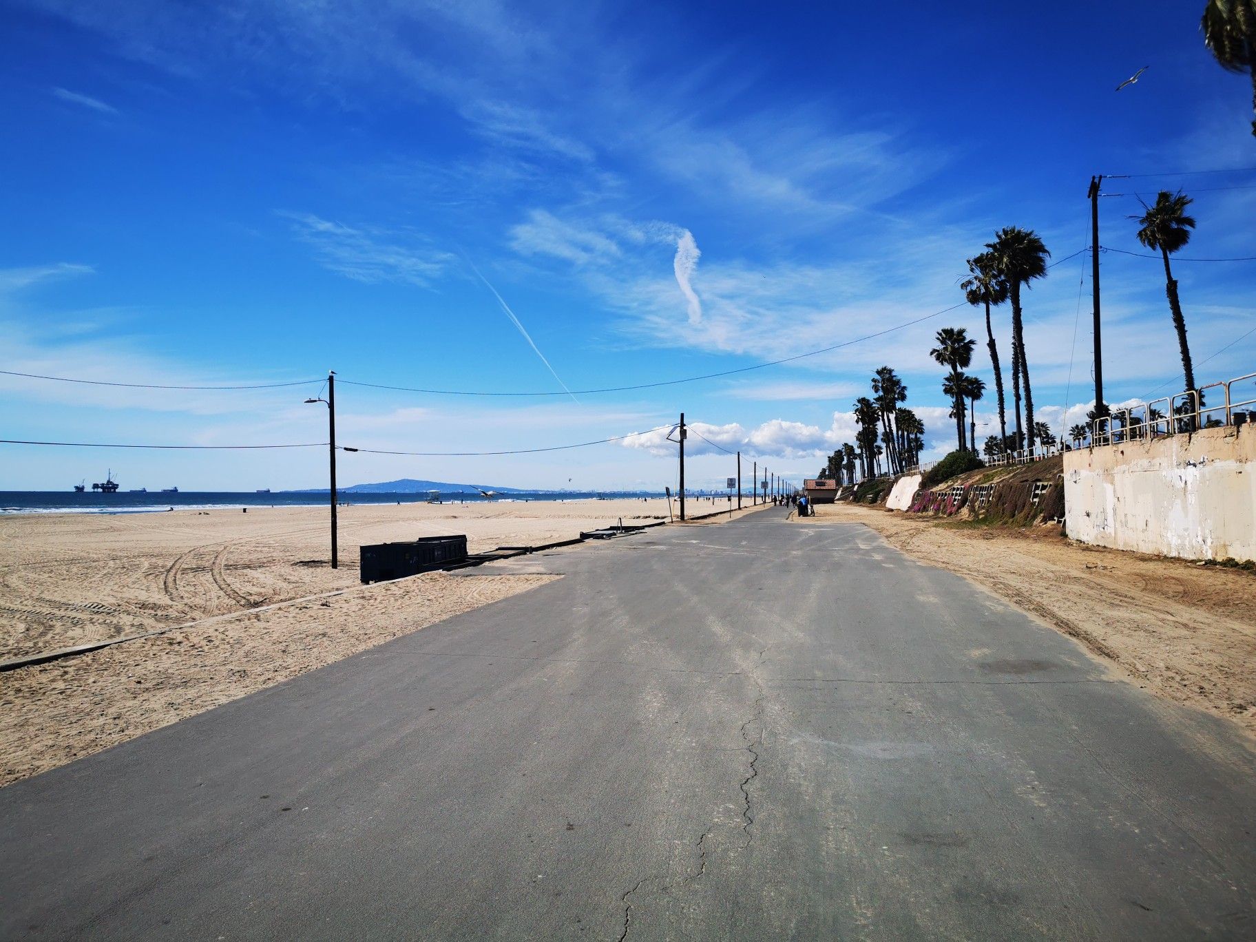 Empty paved road next to a wide sandy beach under a bright blue sky. - Huntington Dog Beach Trail