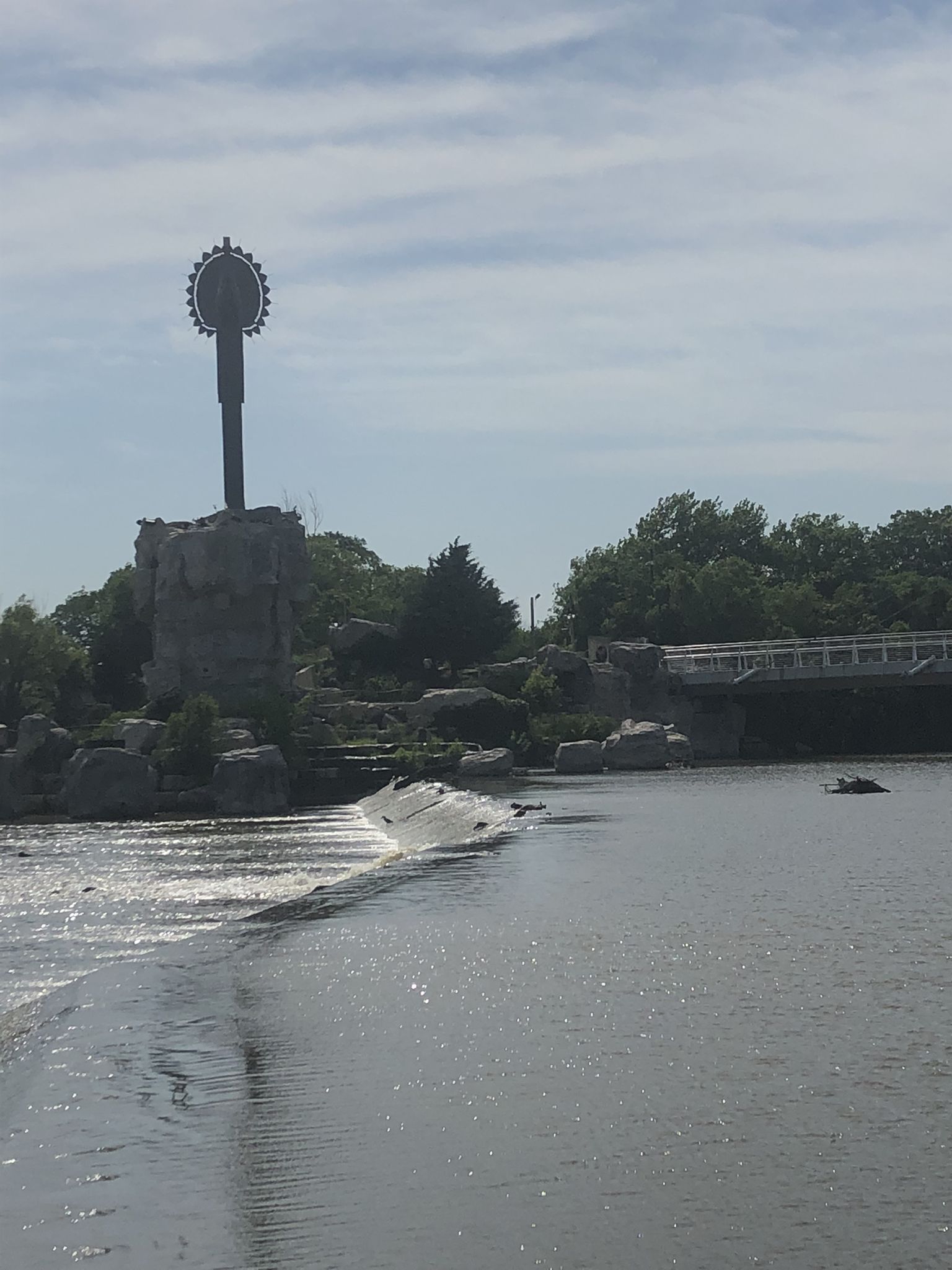 River with a weir, distant sculpture, and bridge under a bright, cloudy sky. - Keeper of the Plains Statue – Keeper of the Plains loop from Wichita