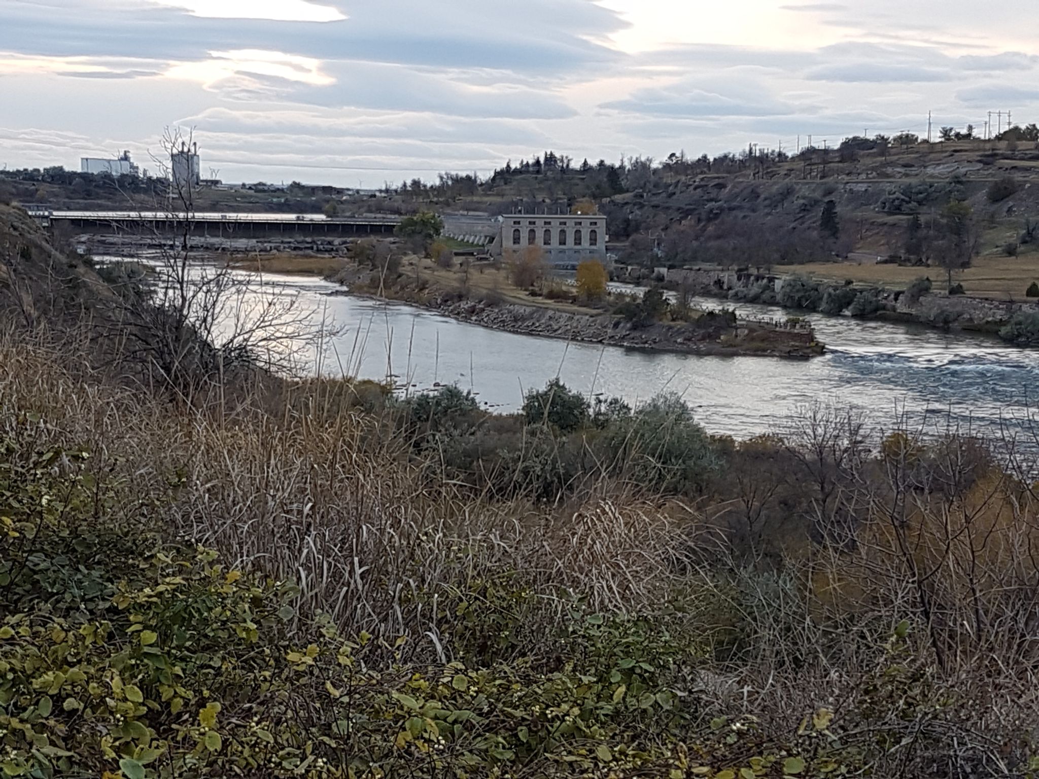 Overcast view of a river flowing past industrial buildings and dry vegetation. - River's Edge Trail