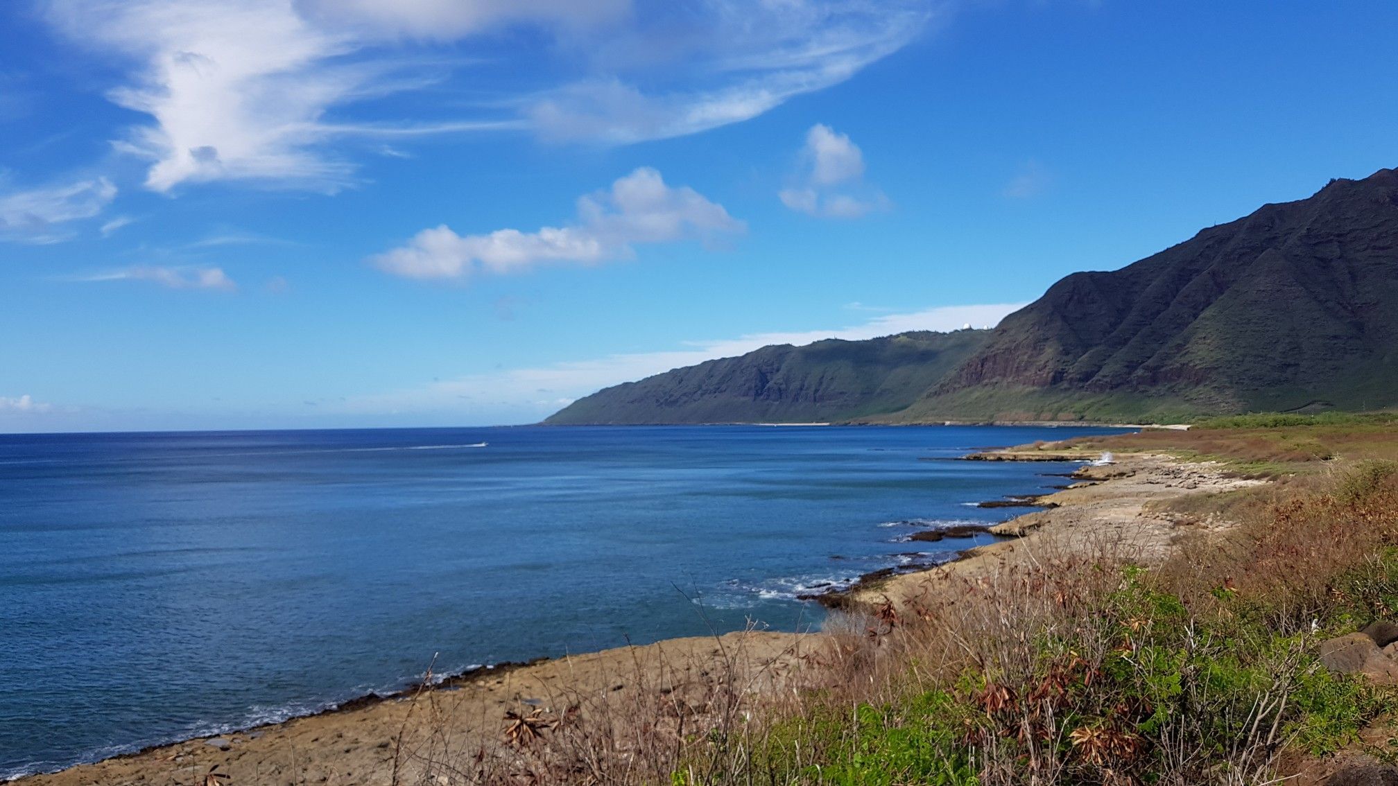 Vast blue ocean meets a rocky coastline with large green mountains under a bright sky. - Kaʻena Point Trail