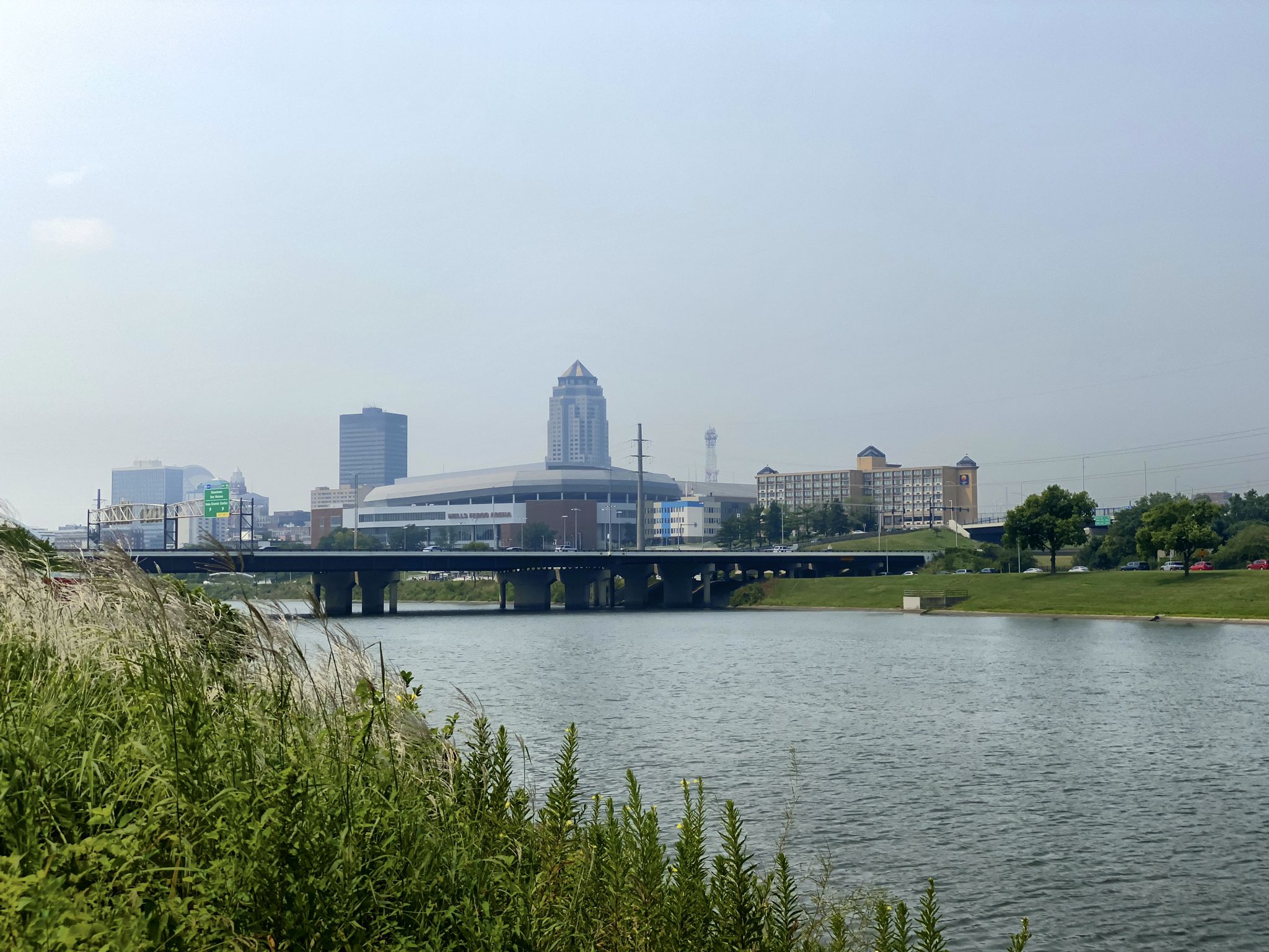 Hazy city skyline with a bridge over a wide river, green foreground. - Bridge Over Des Moines – Des Moines River loop from Johnston