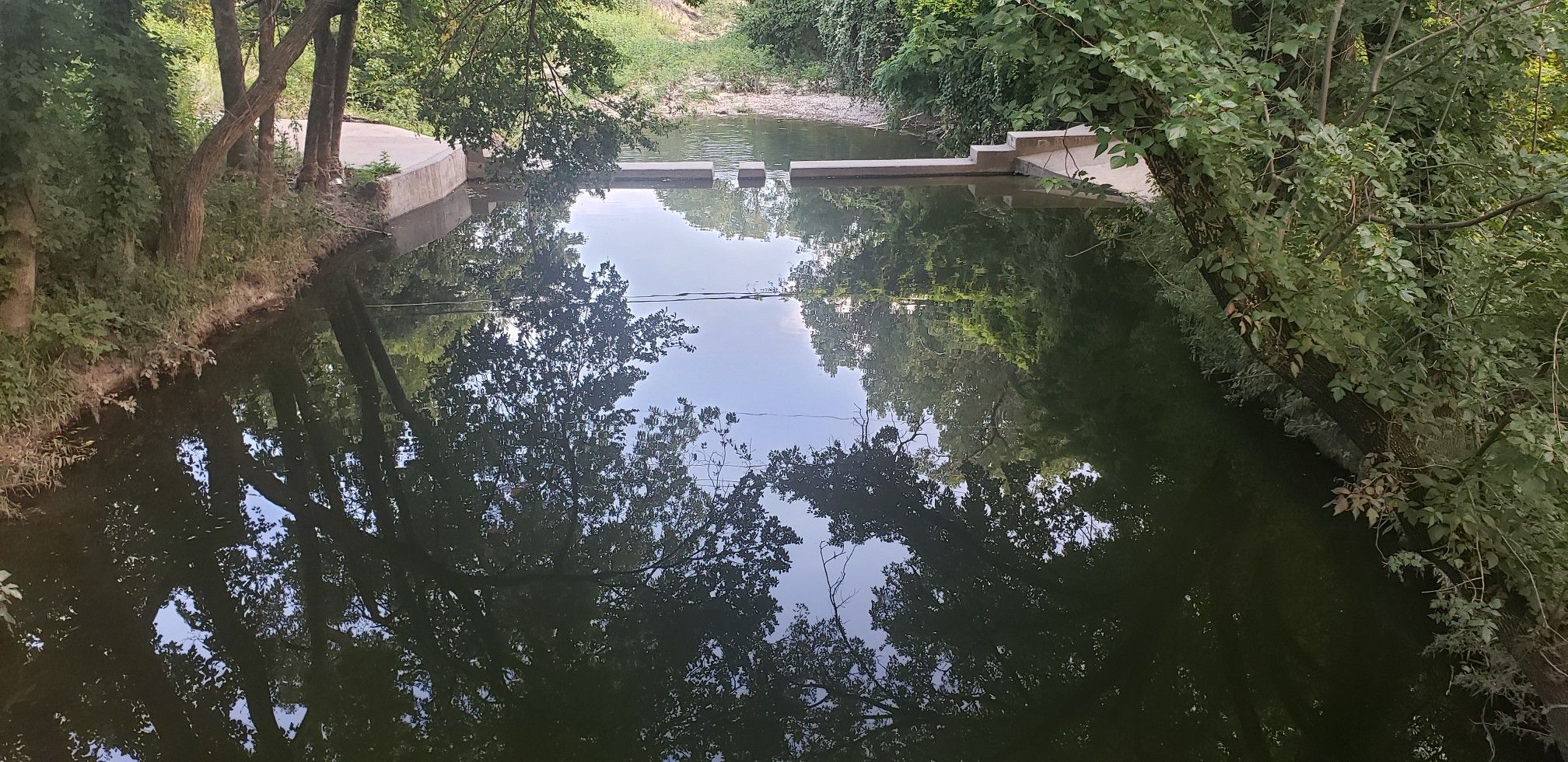 Calm waterway with concrete banks and a small dam, surrounded by dense green trees. - Vitruvian Park and Brookhaven Running Track Loop