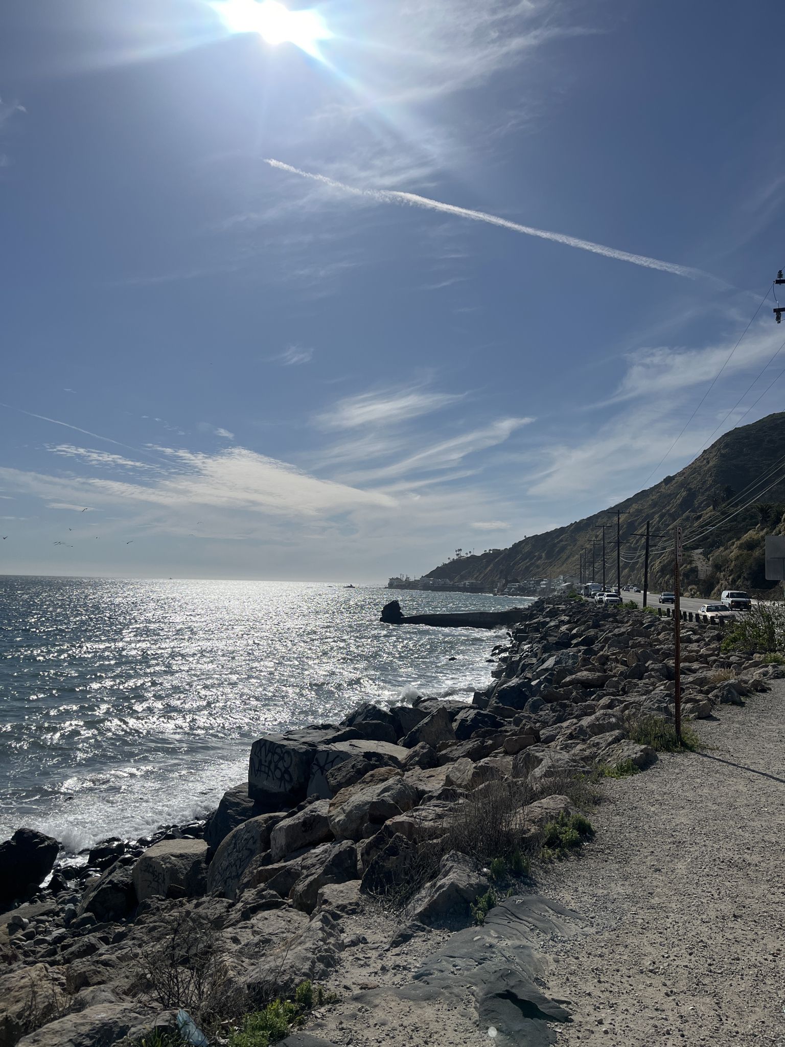 Bright sunny ocean coastline with sparkling water, rocky shore, and distant road. - Piuma Road Loop