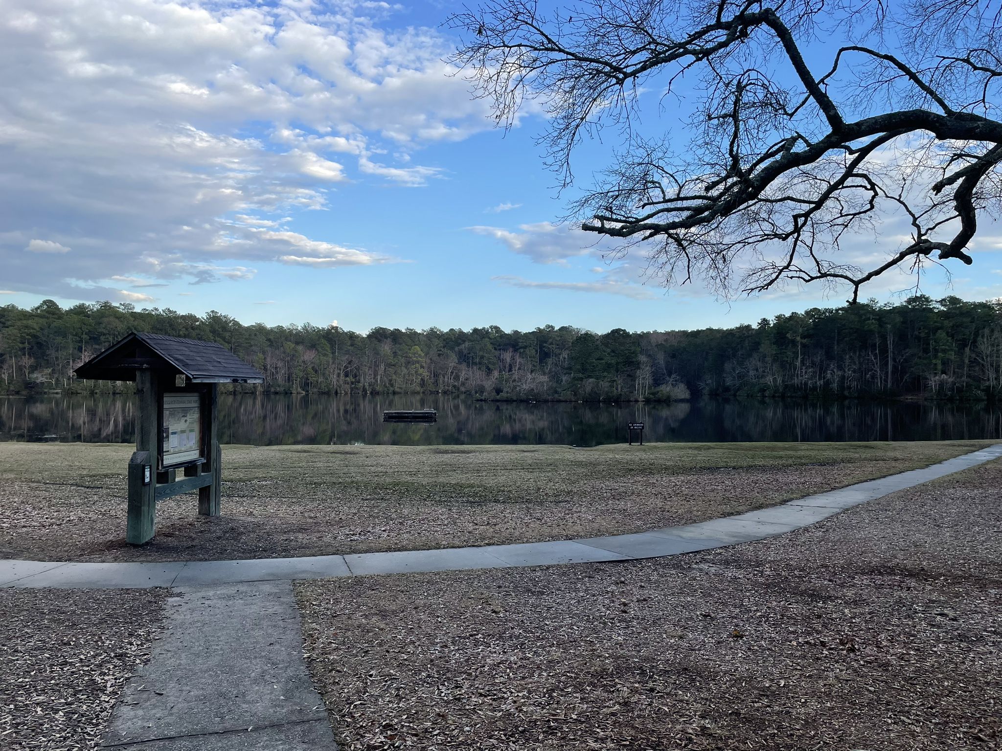 Calm lake, forest, sign, and path under a partly cloudy sky. - Sandhills Hiking Trail