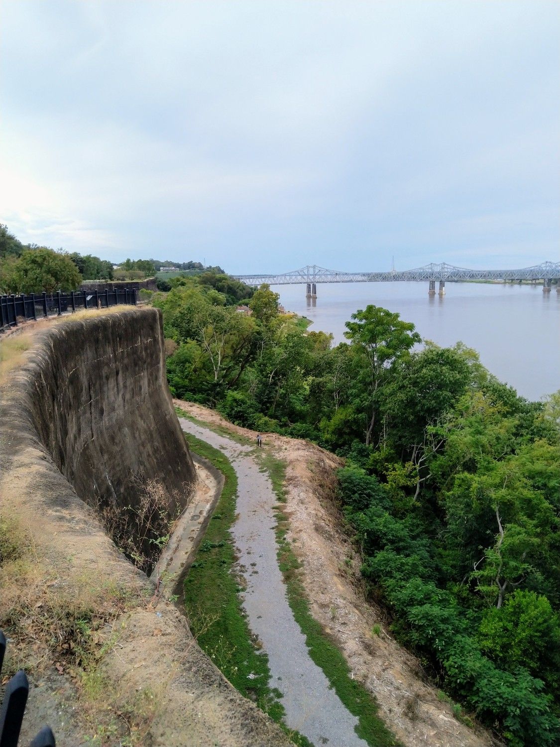 Curved concrete wall beside a winding path, river, and distant bridge. - Natchez Bluff Trail