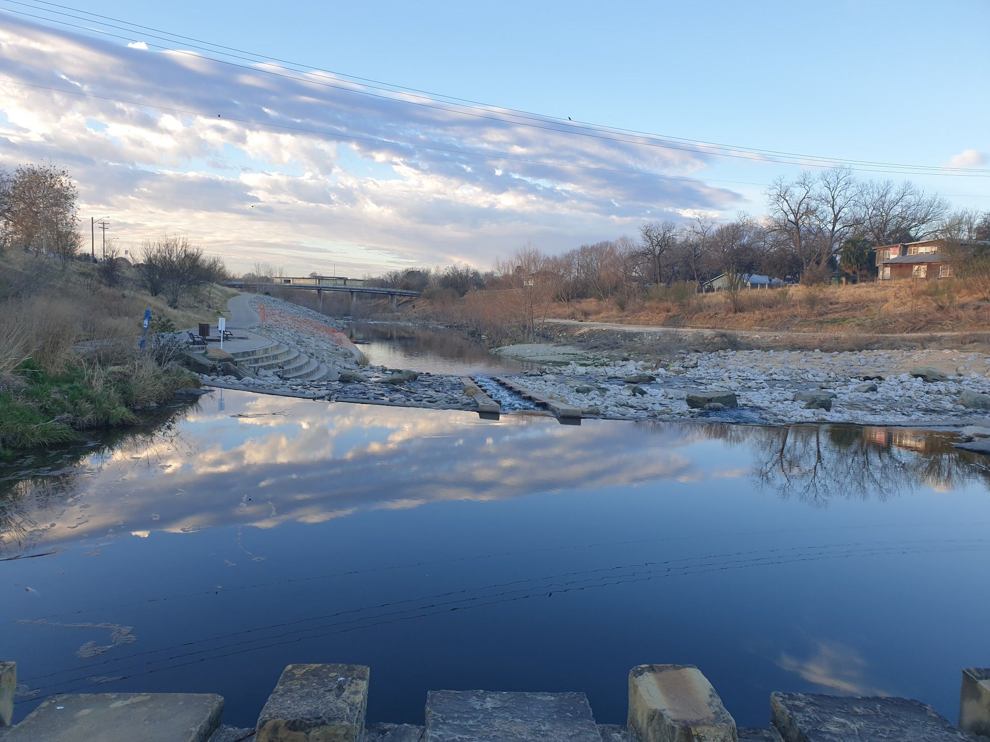 People fishing by a river with rocky rapids under a cloudy sky. - Apache Creek Greenway Loop