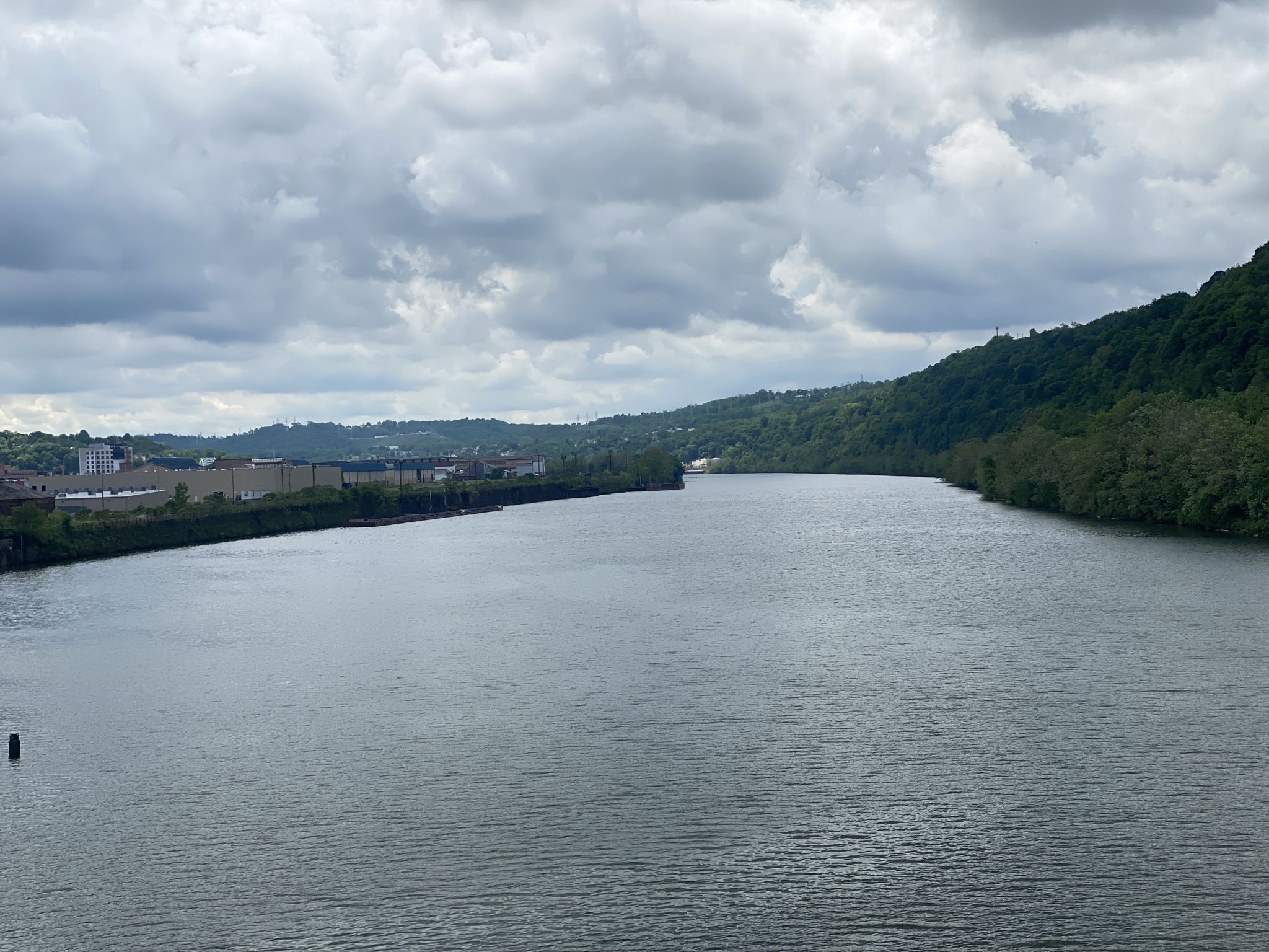Wide river flowing between forested hills and distant industrial buildings under cloudy sky. - Homestead Pump House – View of US Steel Plant loop from Homestead