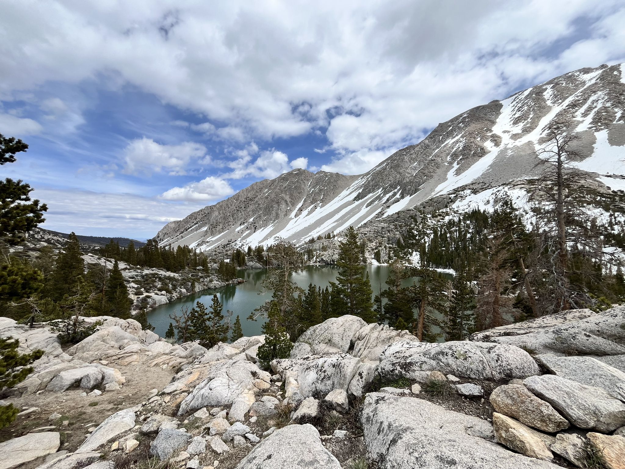 Mountain lake surrounded by snow-capped peaks and pine trees under cloudy sky. - Big Pine Lakes Trail