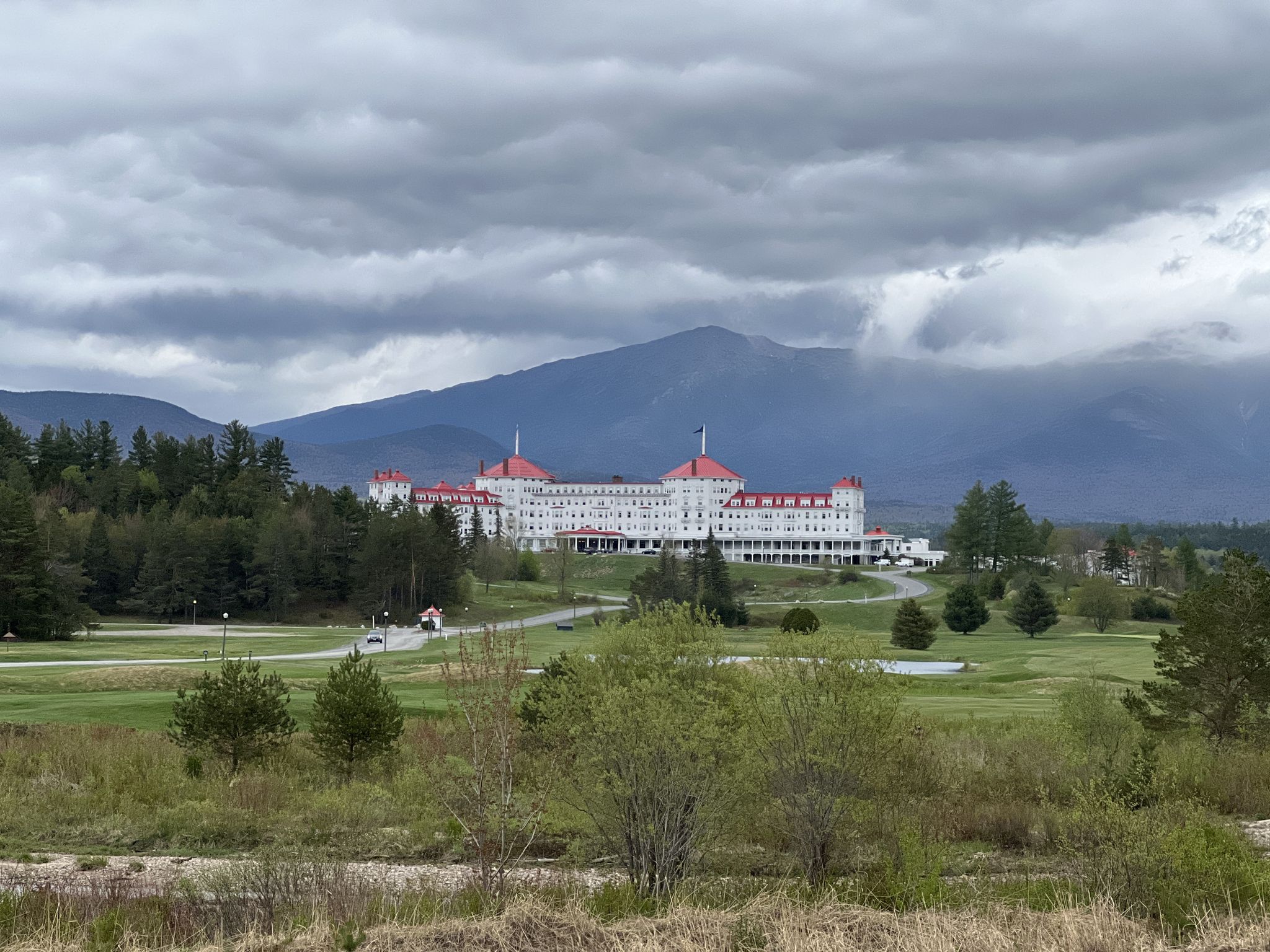 Large white building with red roofs, green fields, and mountains under cloudy sky. - Base Station Road & Mount Clinton Road loop