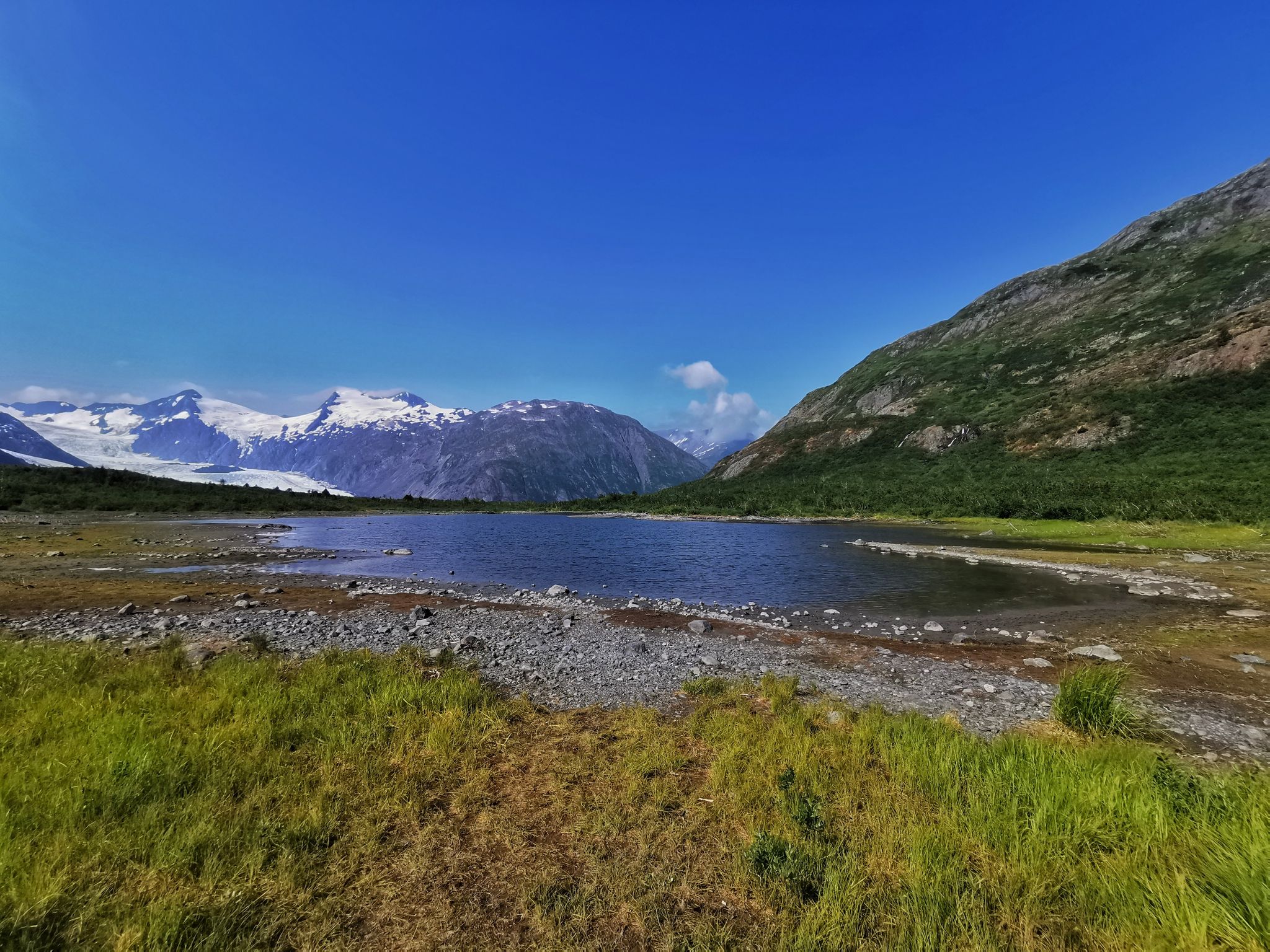 Wide view of a calm lake with rocky shore, surrounded by green and snowy mountains. - Portage Pass Trailhead – Portage Pass loop from Whittier Airport