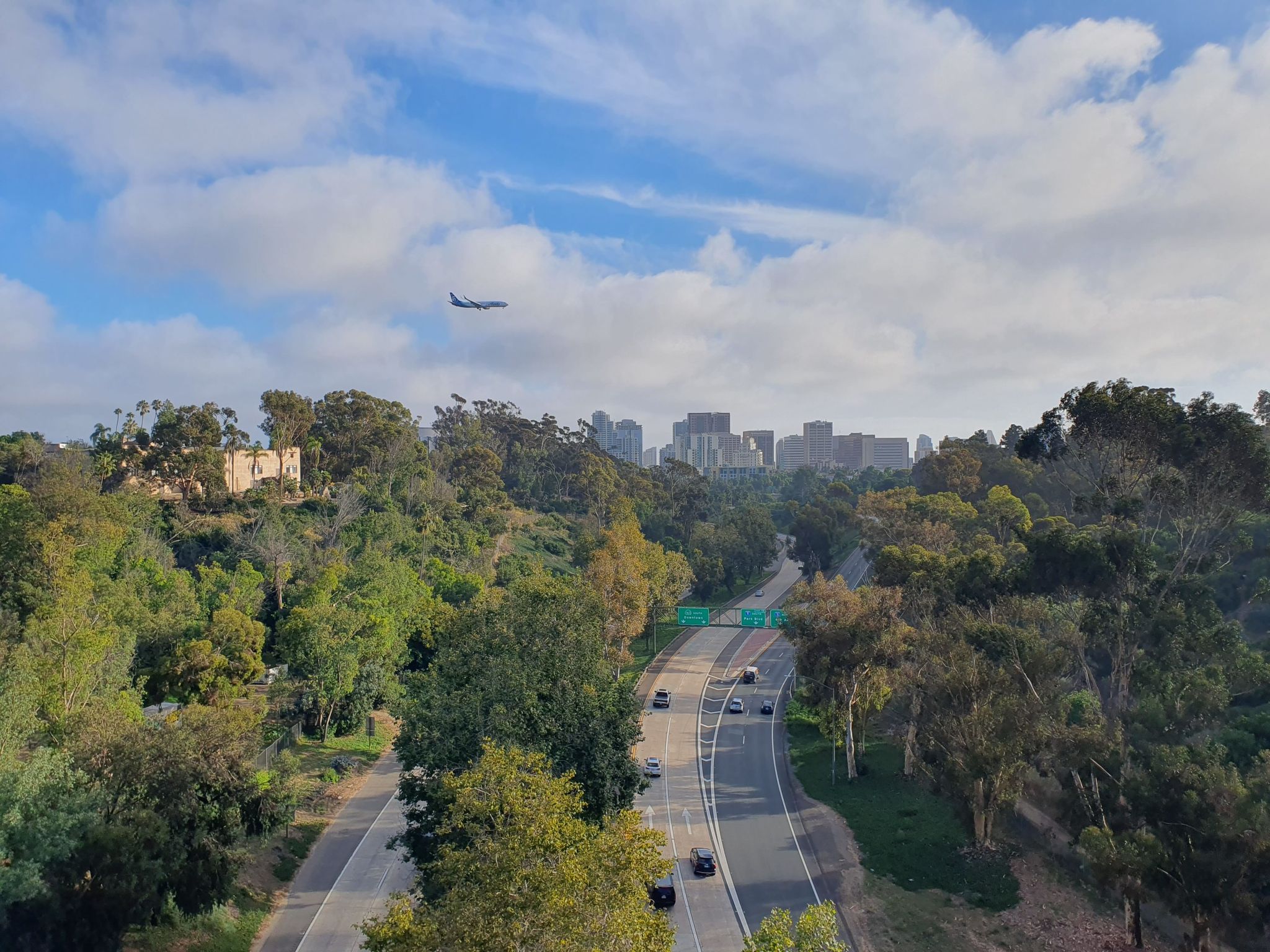 Highway through green hills, a distant city, and an airplane in the sky. - Seven Bridge Walk