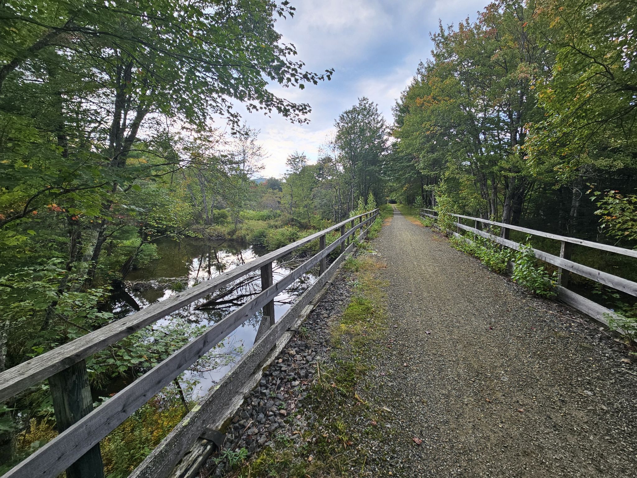 Gravel path with a wooden fence alongside a calm river and green trees. - Northern Rail Trail
