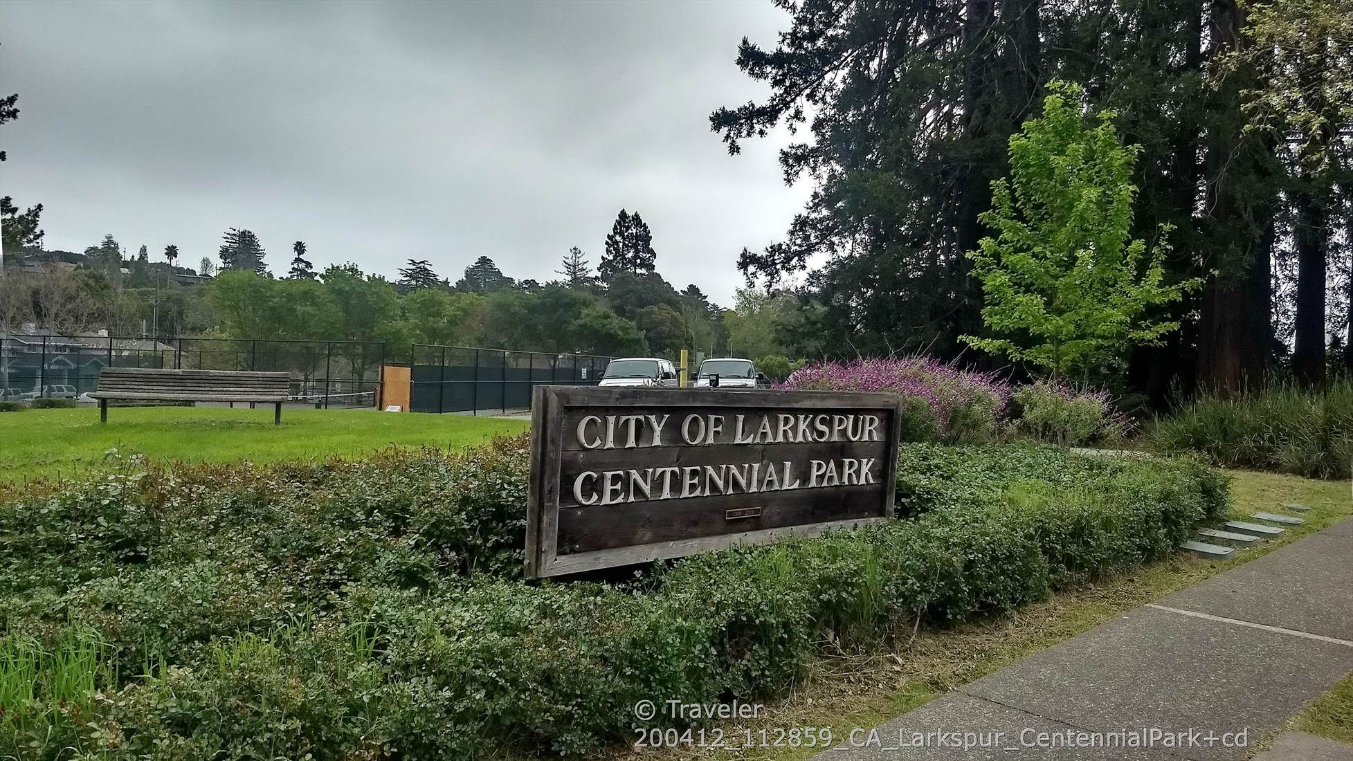 A wooden park sign stands beside a paved path in a grassy, treed area. - Hoo-Koo-E-Koo Loop Trail