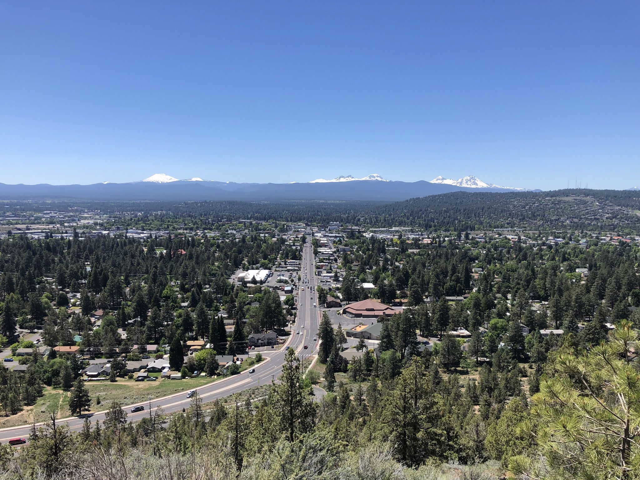 Aerial view of a city, highway, and distant snowy mountains under a clear sky. - Pilot Butte Nature Trail – View of Bend from Pilot Butte loop from Pilot Butte