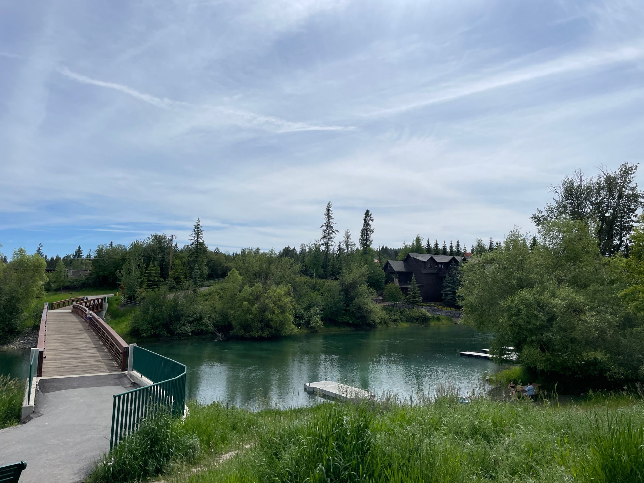 Wooden bridge over a green river, surrounded by lush trees and buildings. - Downtown to Whitefish City Beach via Fish Trails Bike and Pedestrian Path loop