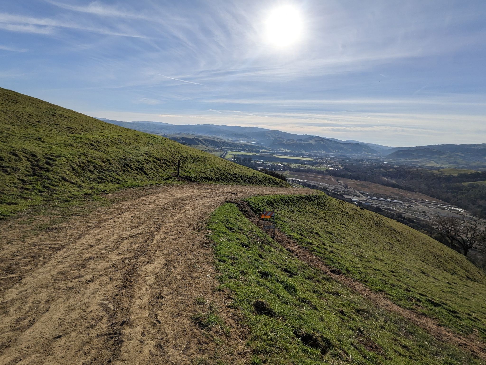 A dirt path curves along a green hillside with a sunny valley view. - Sunol Ridge Trail