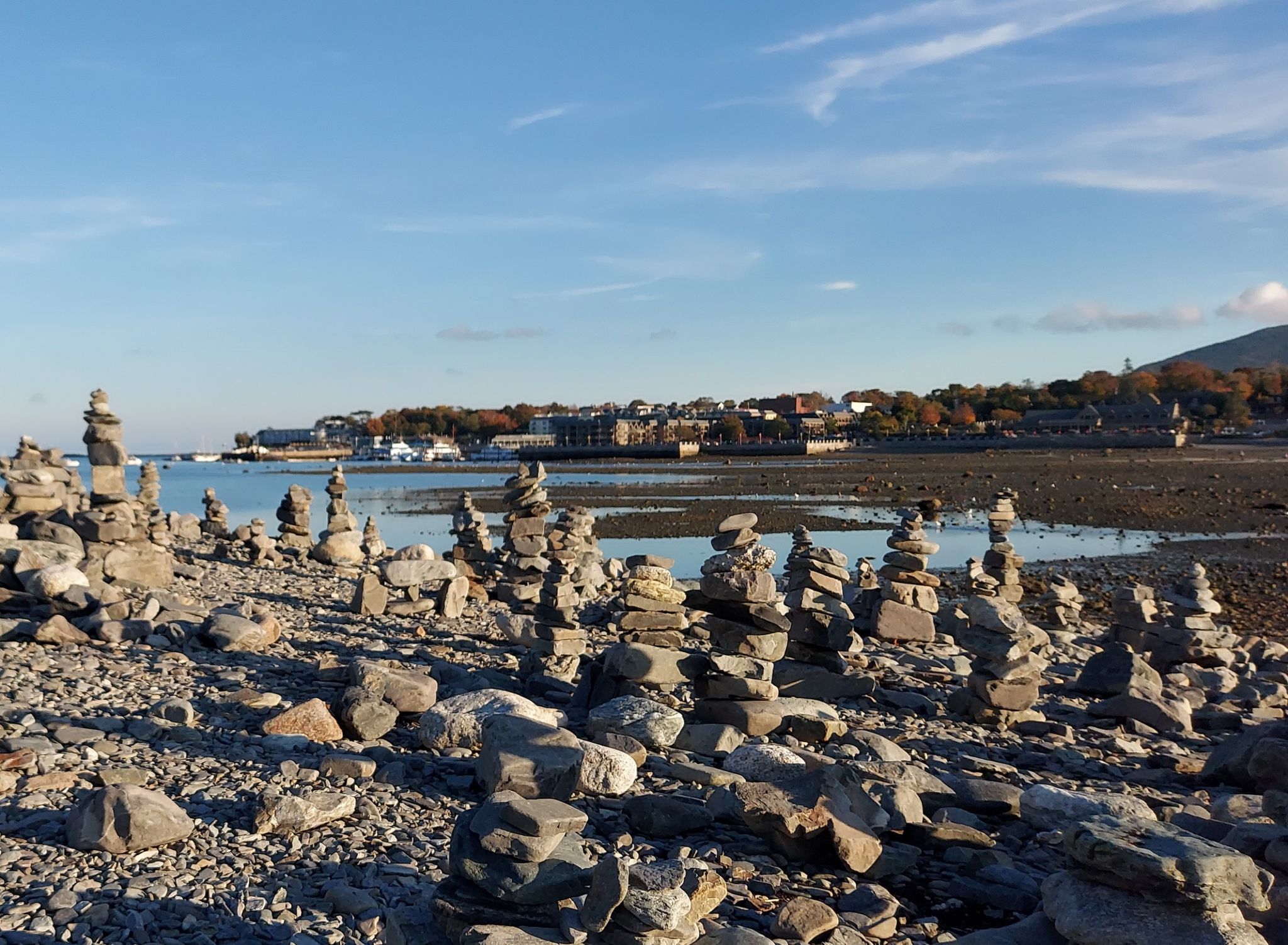 Numerous rock cairns on a rocky shore with water and distant town. - Bar Island Trail