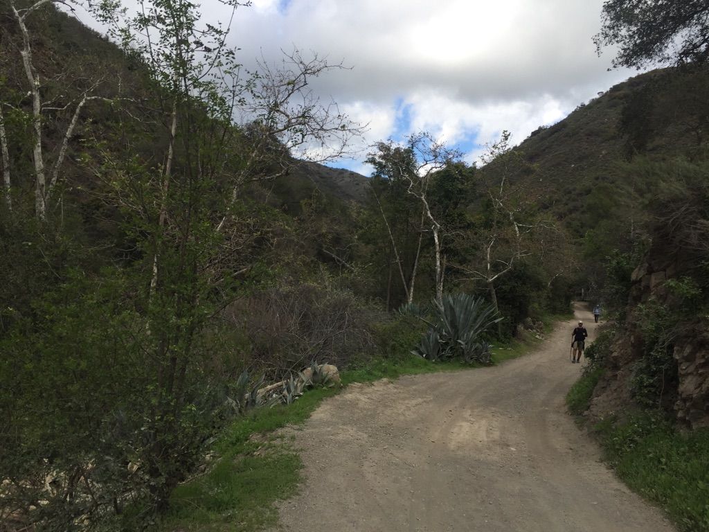 Two people walk on a dirt path through wooded mountains under cloudy skies. - Holy Jim Trail