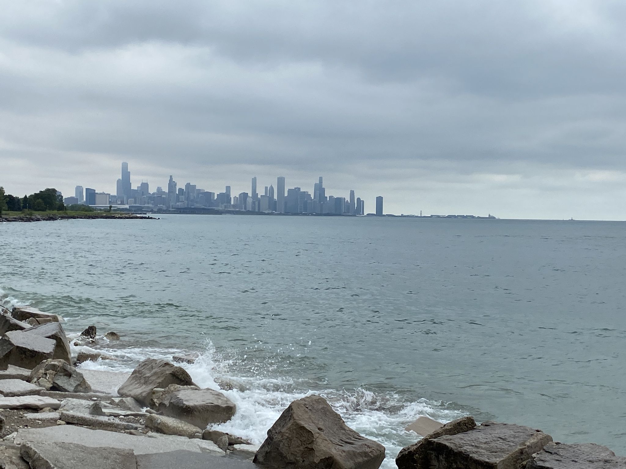 Waves crash on a rocky shore with a distant city skyline under gray clouds. - Museum of Science and Industry – Hyde Park loop from 63rd Street