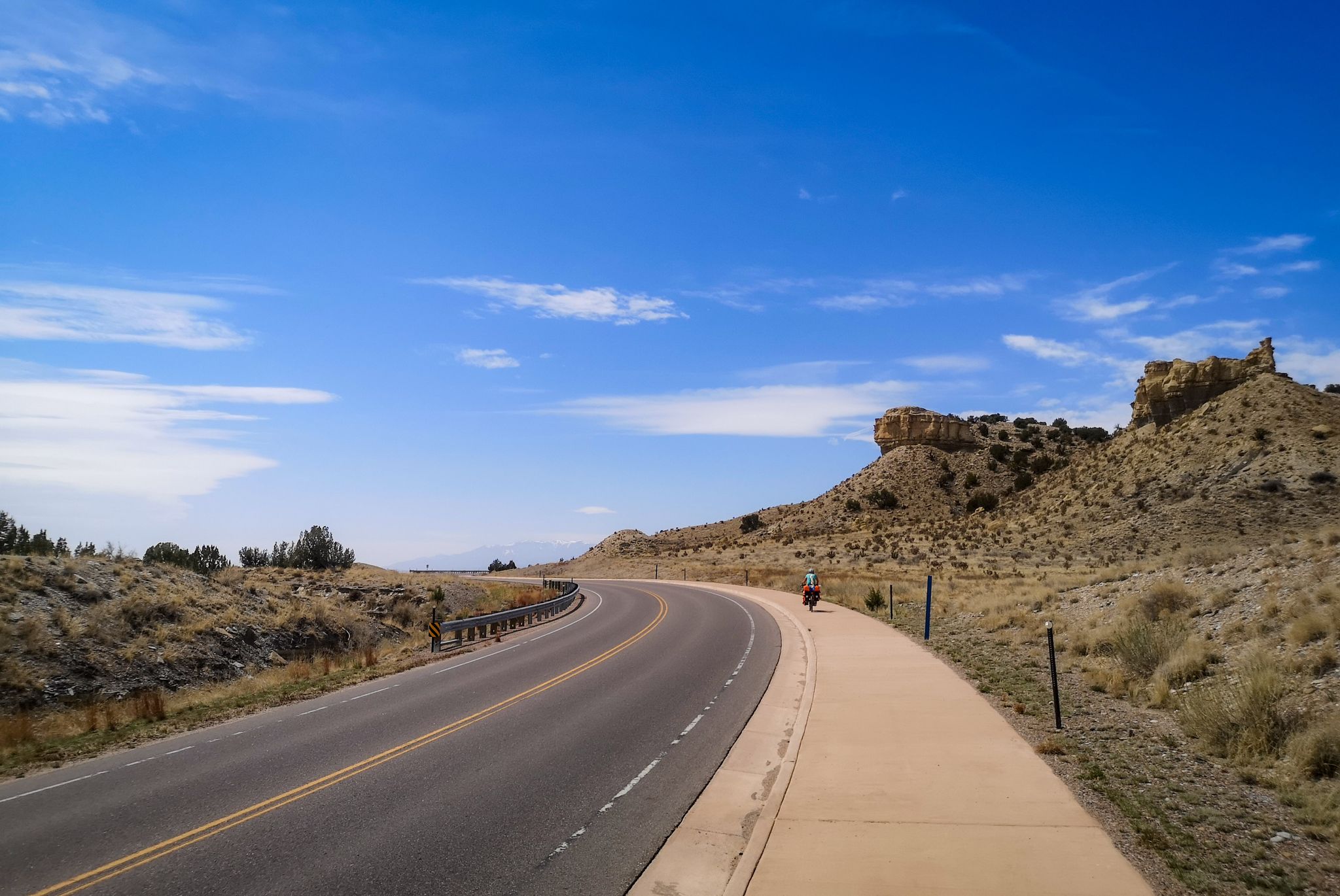 Curving road and path through arid hills with a distant cyclist. - View of Pueblo Reservoir Dam – South Shore Trail Network loop from Pueblo West