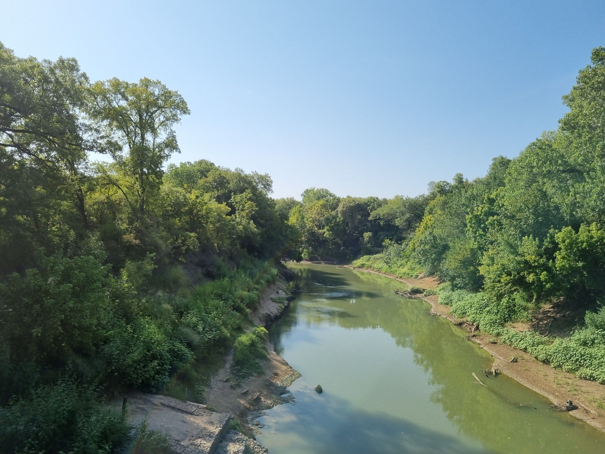 Calm river flowing through a lush, green wooded valley under a clear blue sky. - River Legacy Trail