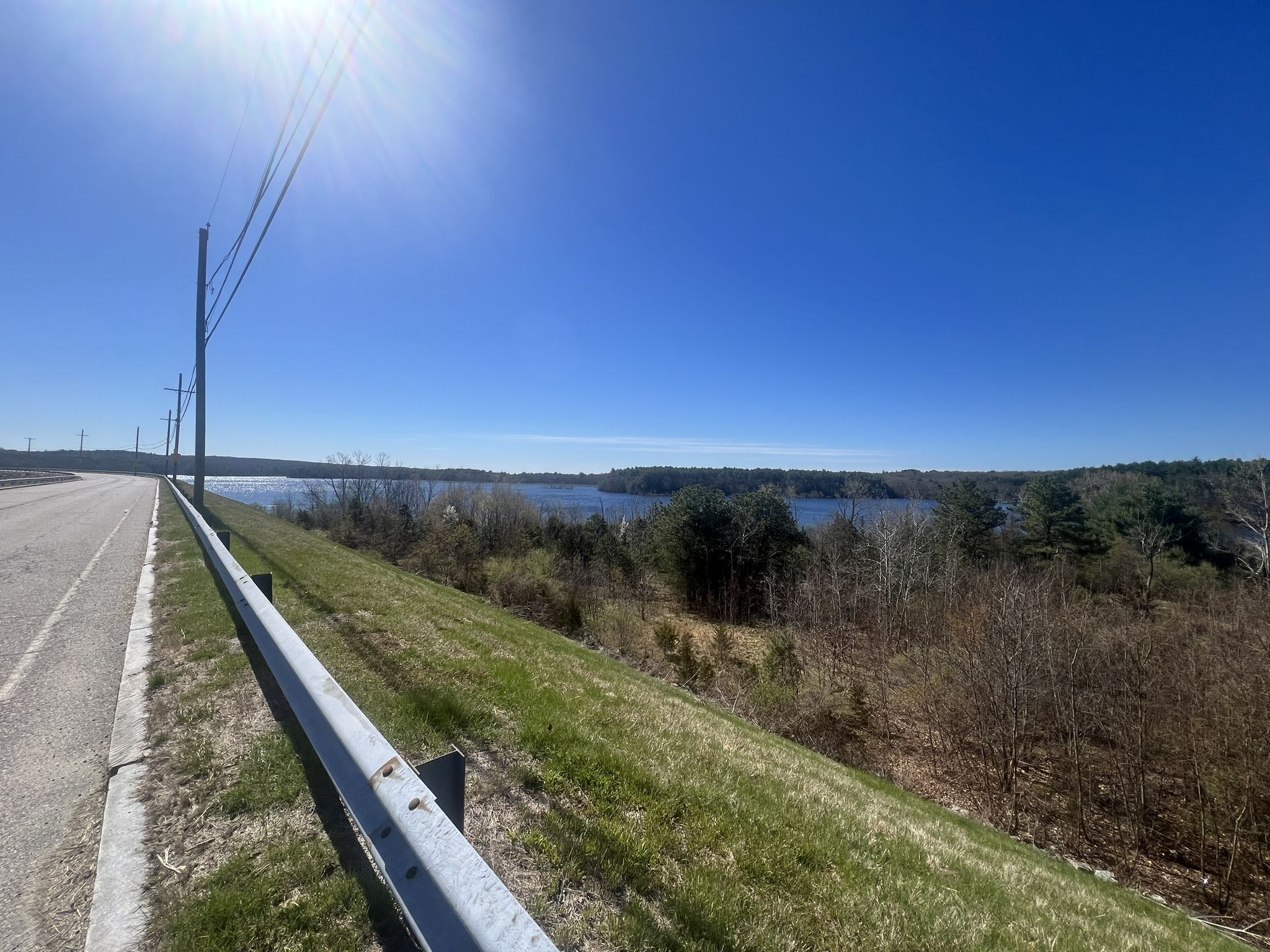 A sunny road with a guardrail overlooking a large lake and distant trees. - Massachusetts to Rhode Island Loop