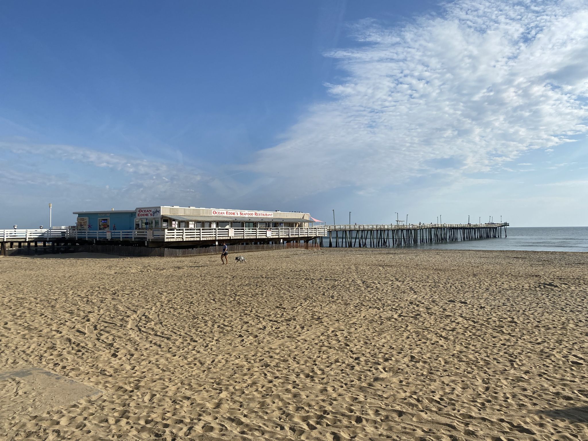 Wide sandy beach with a long pier and building under a partly cloudy sky. - Virginia Beach Boardwalk