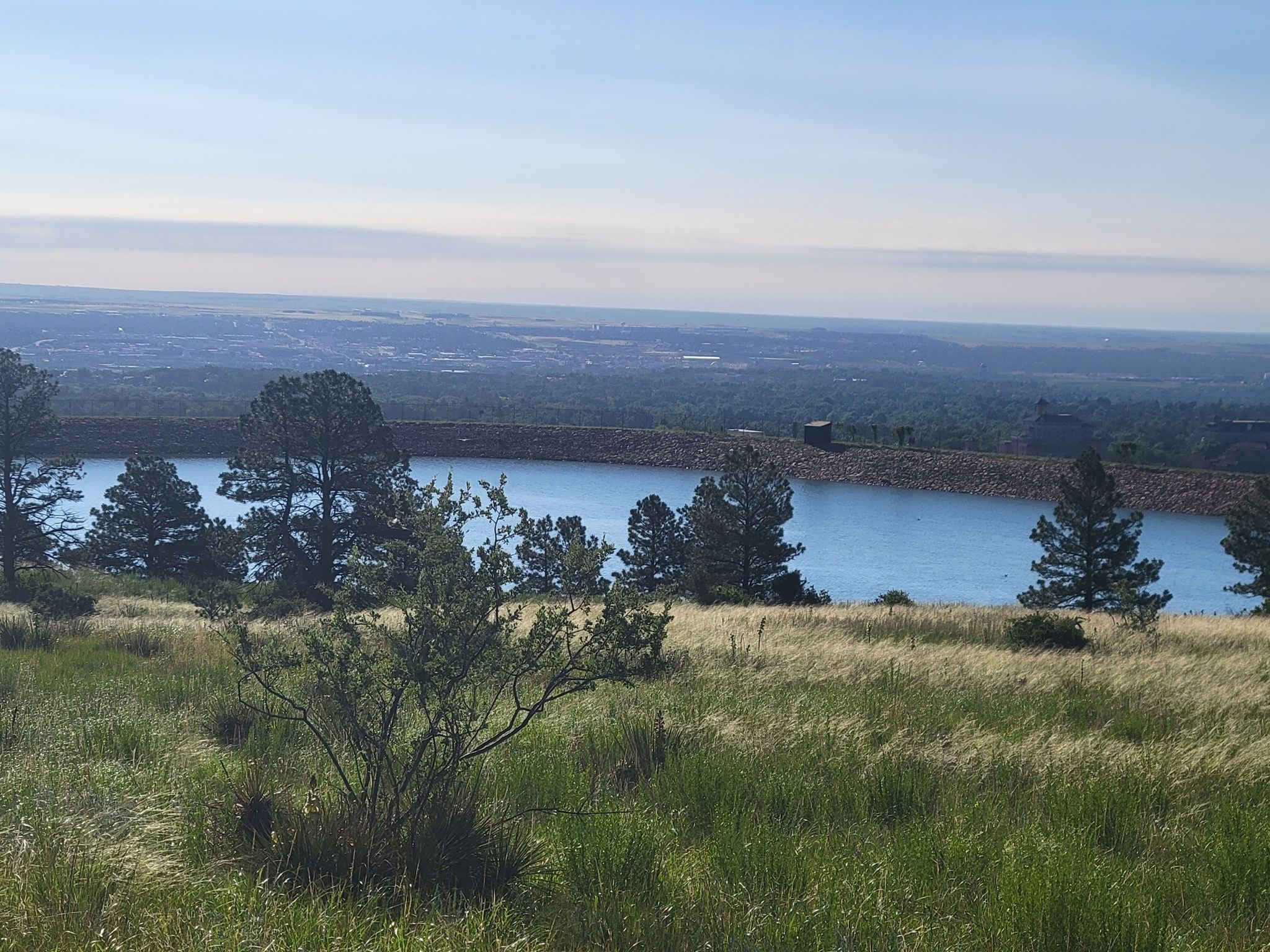 Sunny view of a reservoir surrounded by trees, with a distant city. - Seven Bridges Trail – Viewpoint on Gold Camp Road loop from The Broadmoor