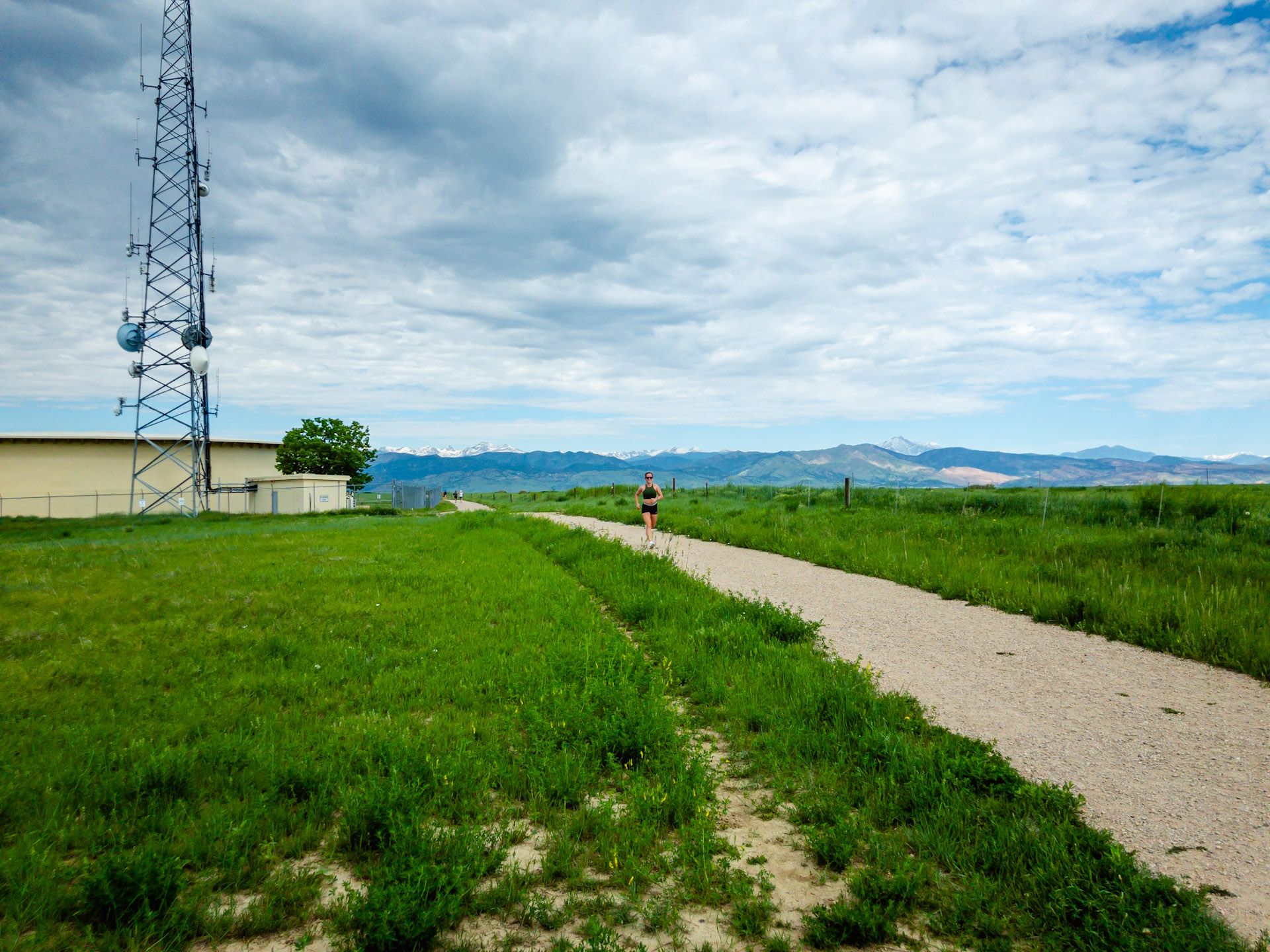 Person running on a dirt path next to a tall tower under a cloudy sky. - East Boulder - Gunbarrel Loop
