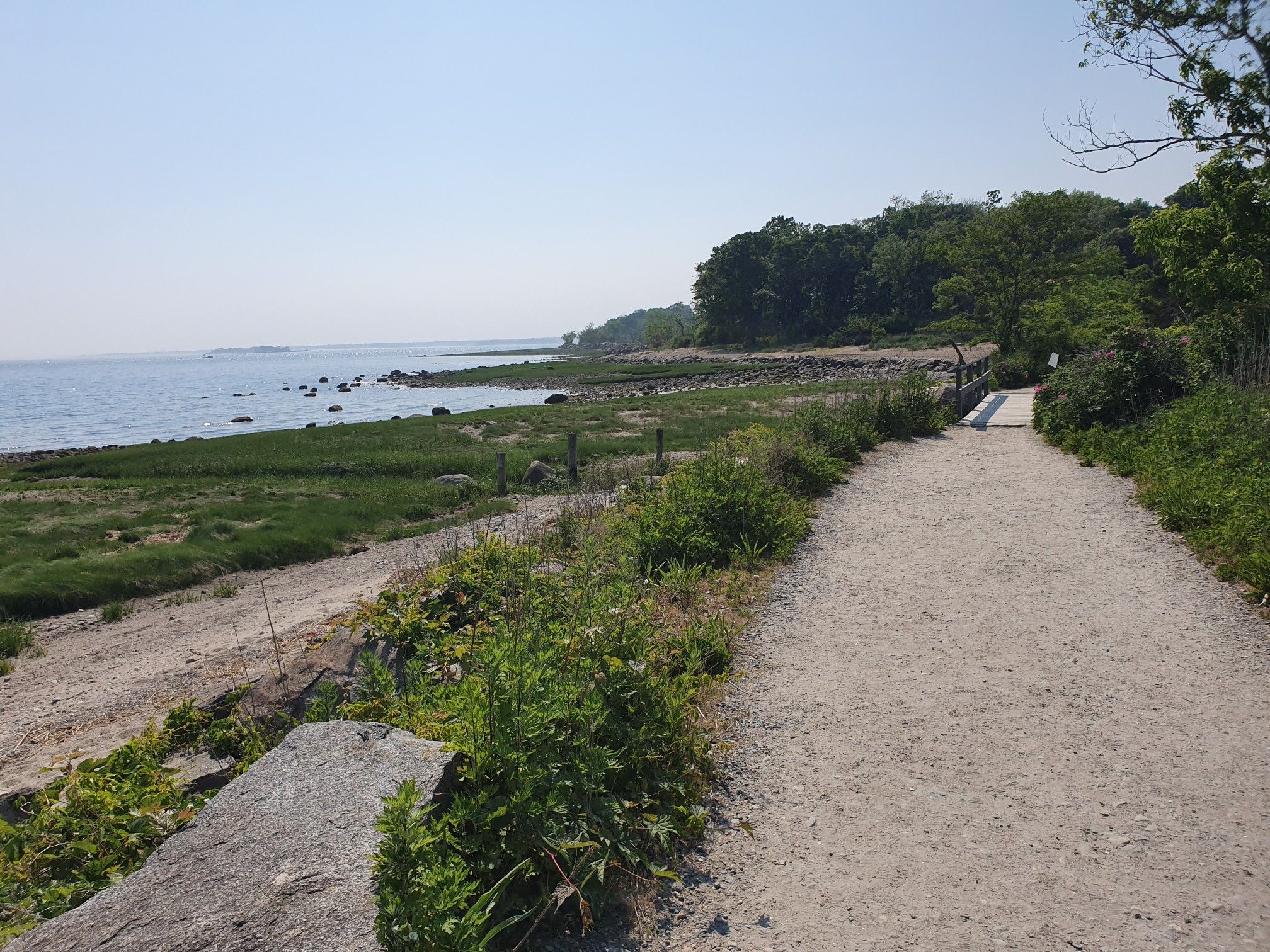 A dirt path runs alongside a rocky shoreline and calm water on a sunny day. - Greenwich Point Park Loop