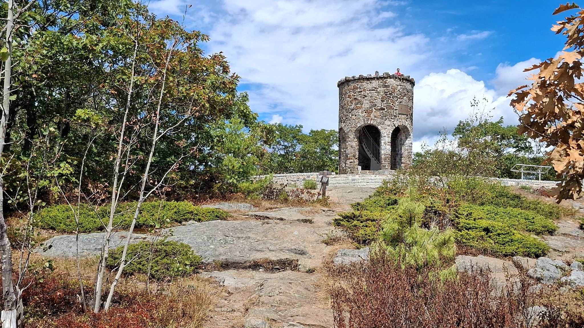 Stone tower on a rocky hill surrounded by trees under a cloudy sky. - Mount Battie Trail