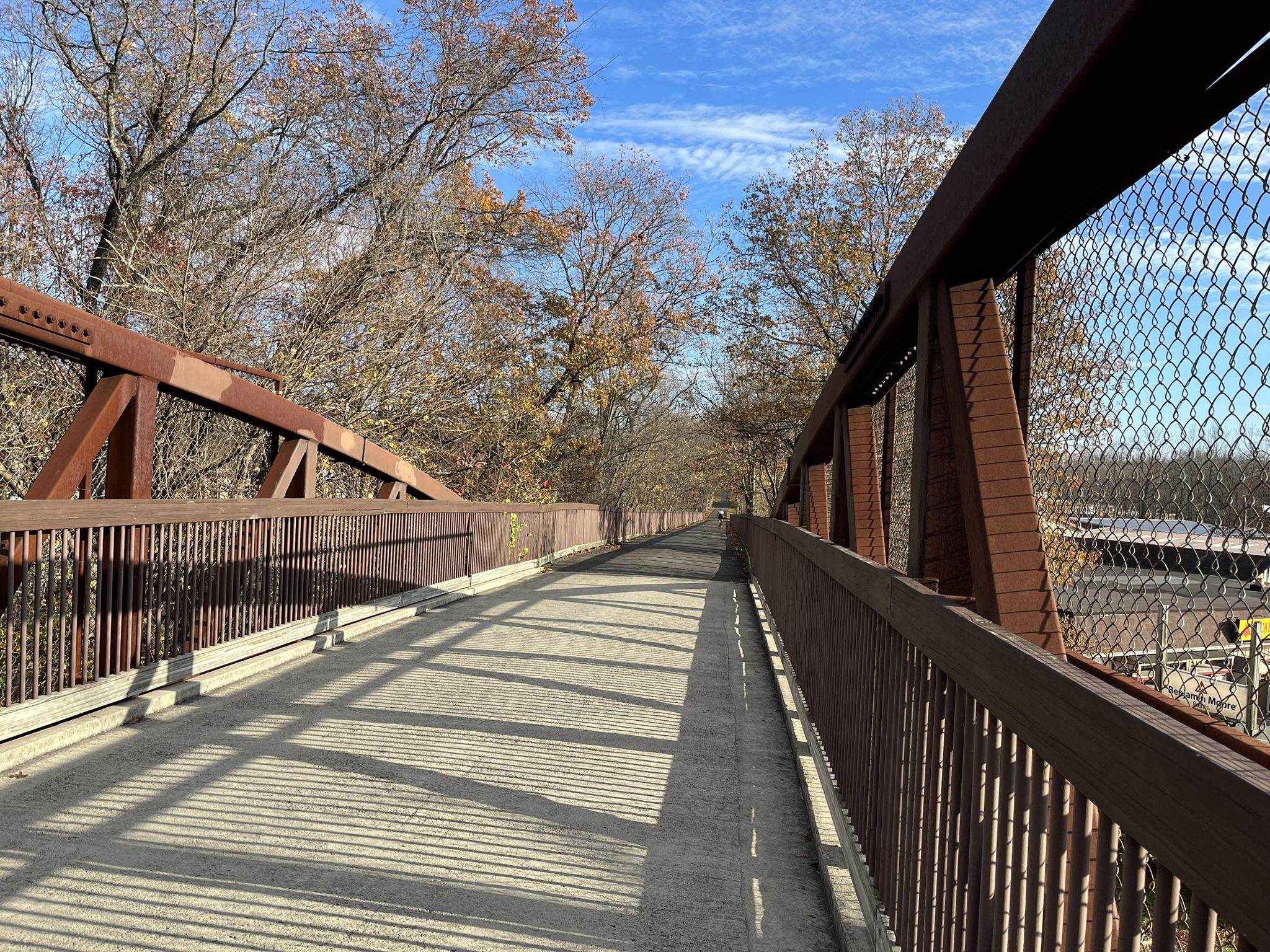 Pedestrian bridge with metal trusses and chain-link fences, surrounded by autumn trees. - Joseph B. Clarke Rail-Trail
