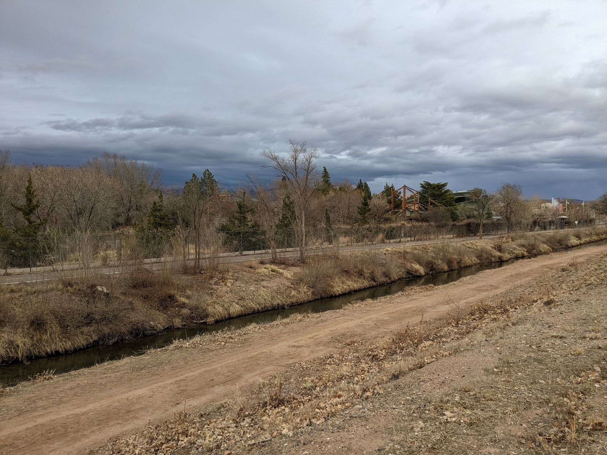 A narrow canal and dirt path under a cloudy sky with bare trees. - View of Rio Grande – Paseo del Bosque Trail loop from Old Town