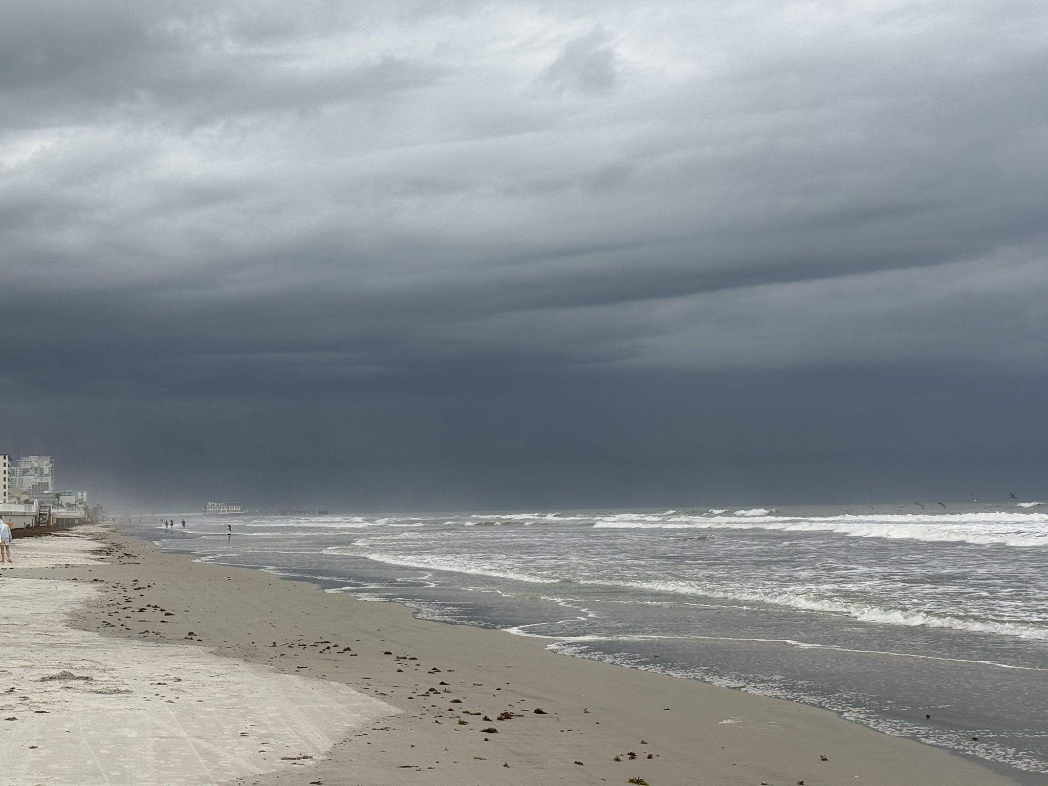 Dark, stormy clouds hang over a beach with crashing waves and distant figures. - Daytona Beach
